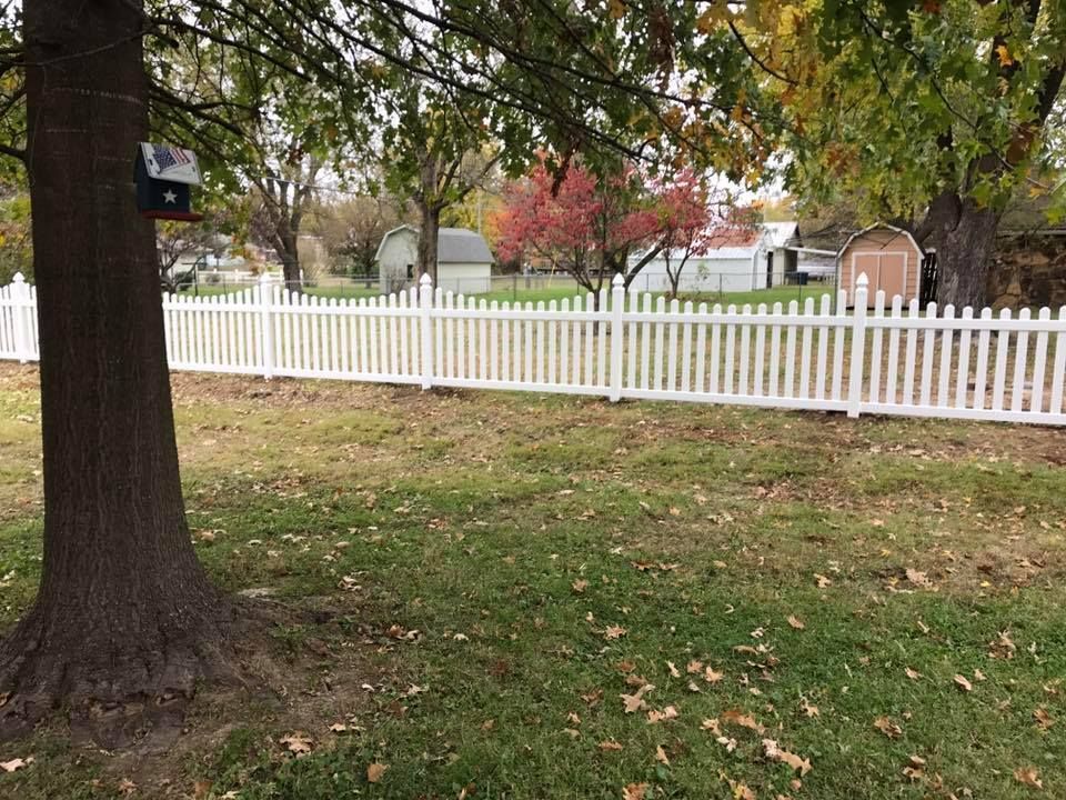 A white picket fence runs across a grassy, tree-filled yard with a birdhouse mounted on a large tree in the foreground.