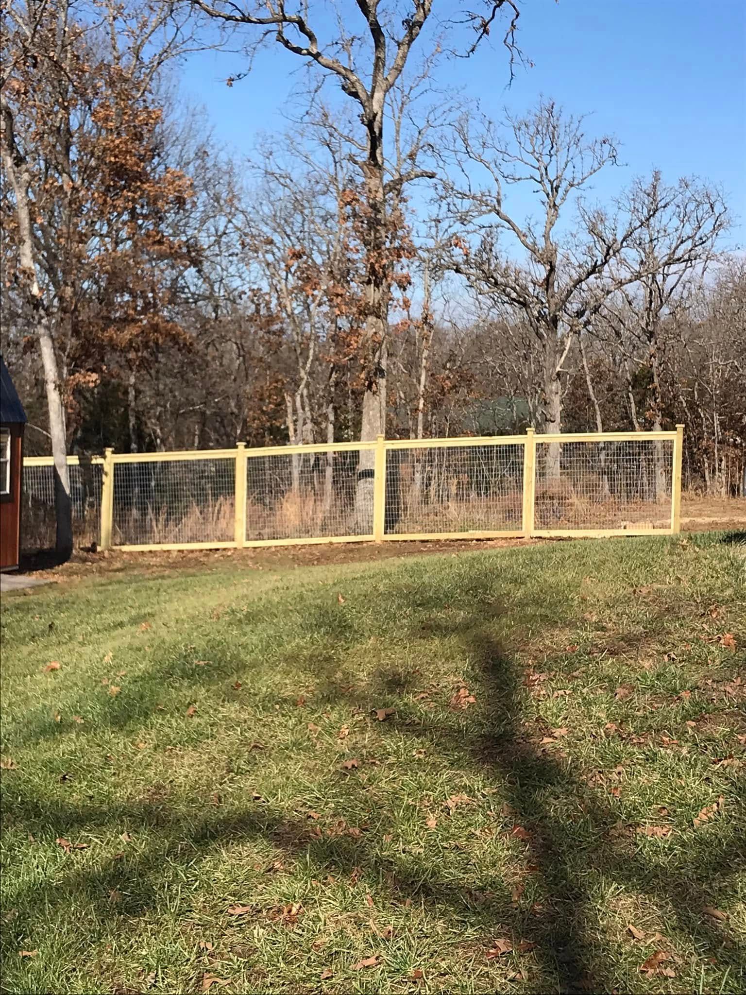 A new wooden fence with wire mesh in a grassy yard, with a treeline and a blue sky in the background.