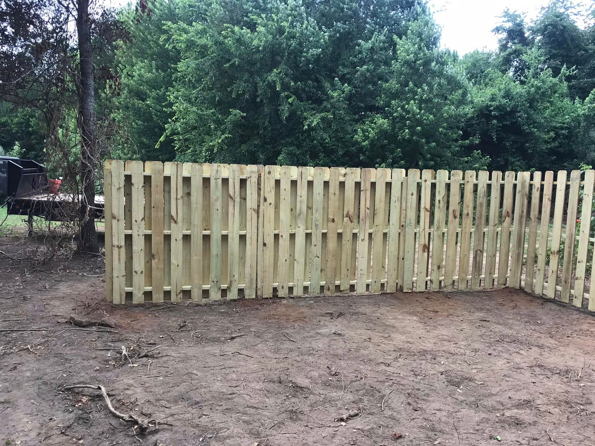 A light-colored wooden picket fence stands in a dirt yard in front of green trees.