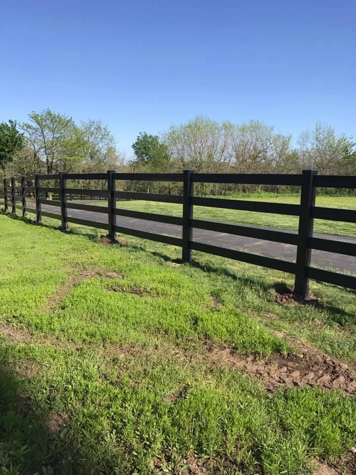 A black four-rail wooden fence runs diagonally across a lush green grassy field under a clear blue sky.