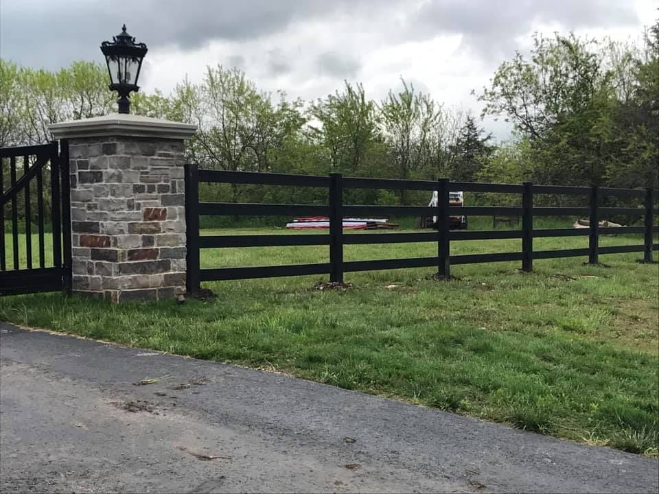 A black, four-rail wooden fence extends from a stone pillar topped with a decorative lamp, set against trees and sky.