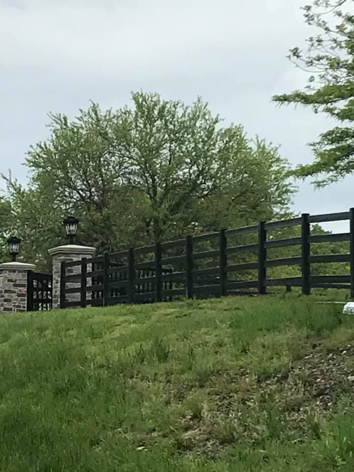A black four-rail fence with brick posts topped with lamps sits on a grassy slope before a large tree under a gray sky.