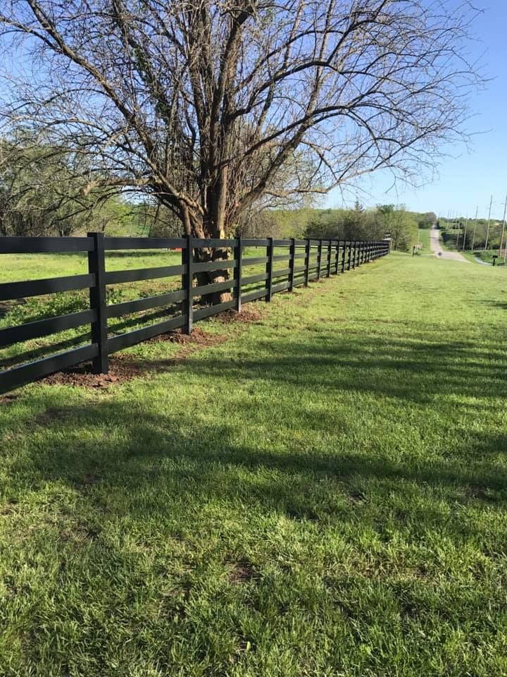 A long black wooden fence runs through a grassy field toward a tree and a road in the distance under a blue sky.