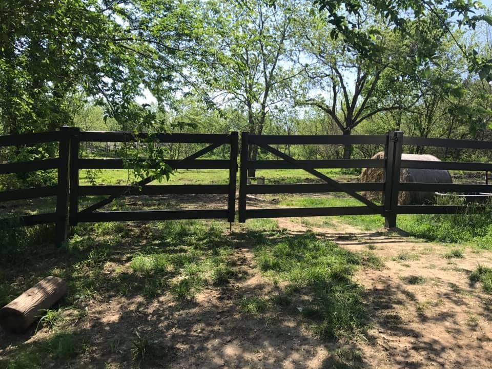 A split wooden farm gate stands closed in a grassy, wooded field on a sunny day.