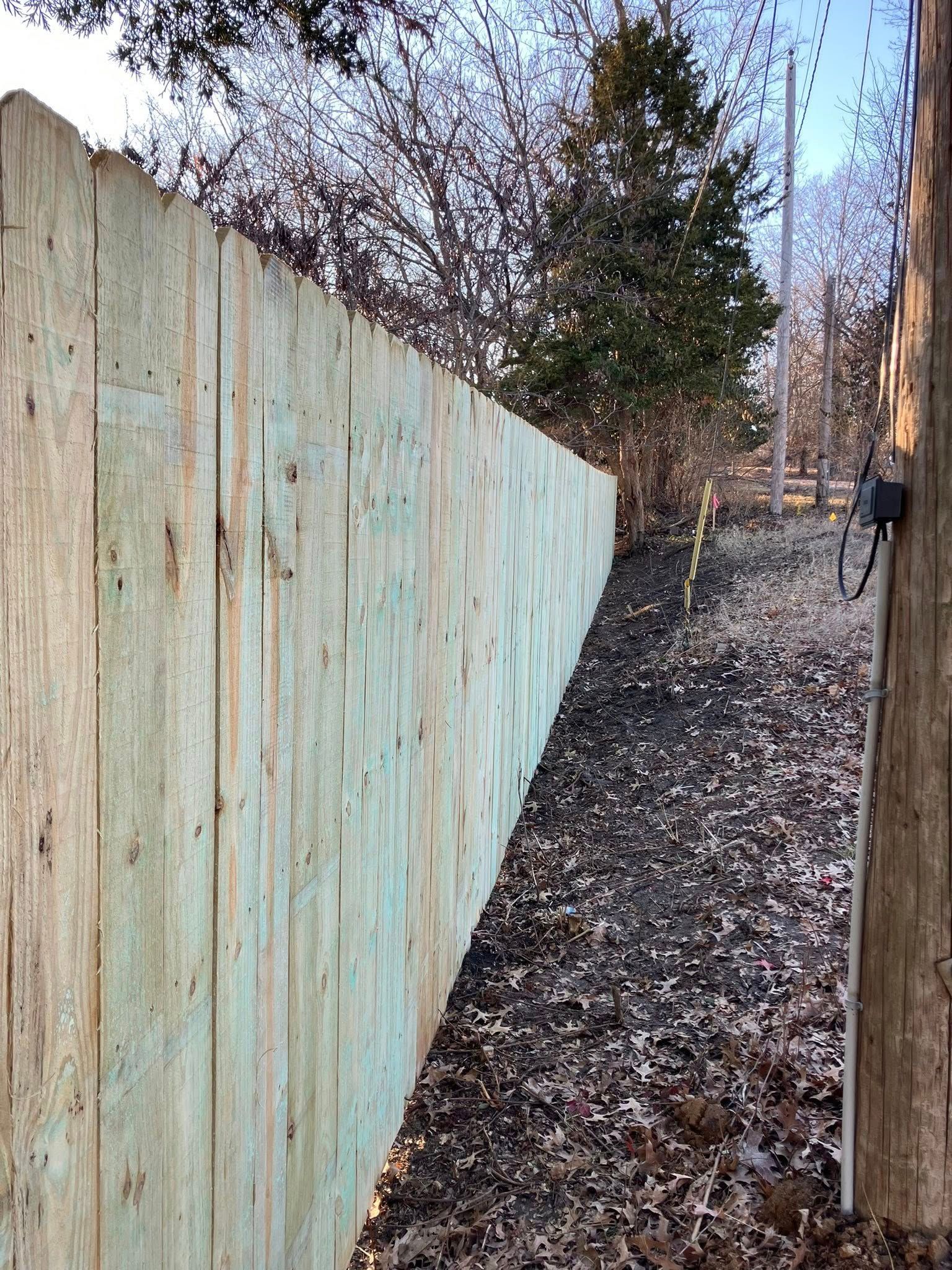A new wooden privacy fence runs along a gravel path covered in fallen leaves under trees.