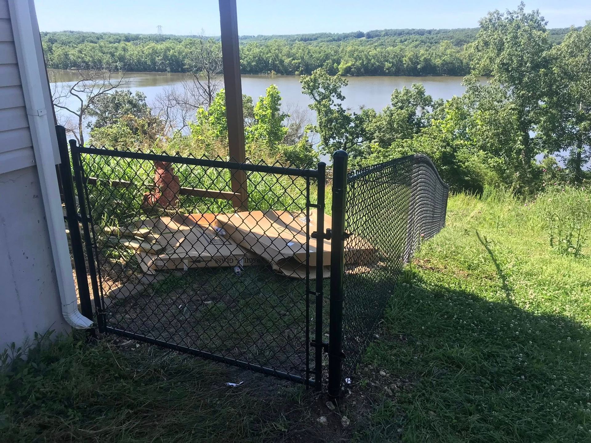 A black chain-link fence gate stands on a grassy hill overlooking a river and treeline on a sunny day.