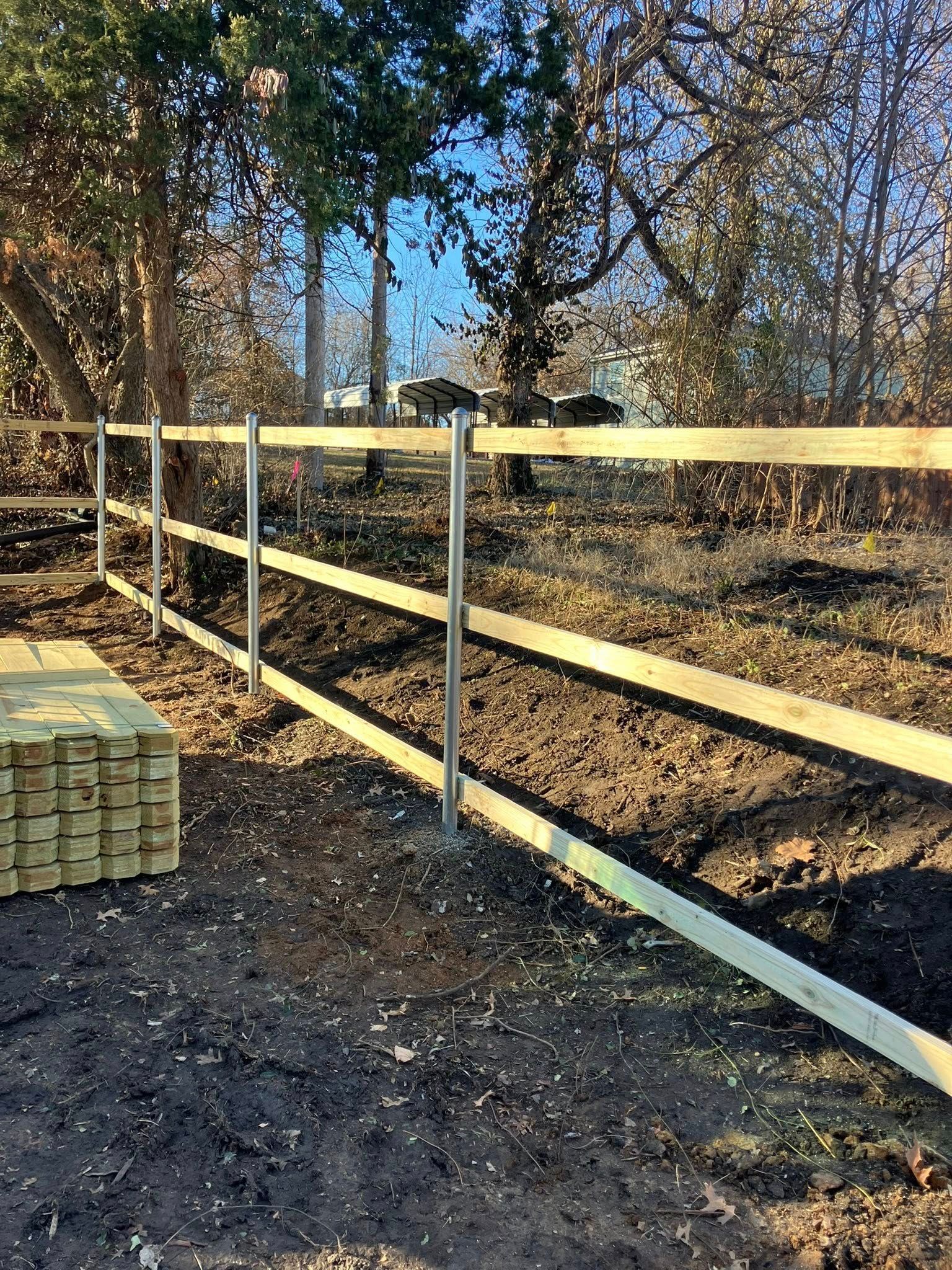A three-rail wooden fence under construction, with a stack of lumber nearby on dark dirt ground.