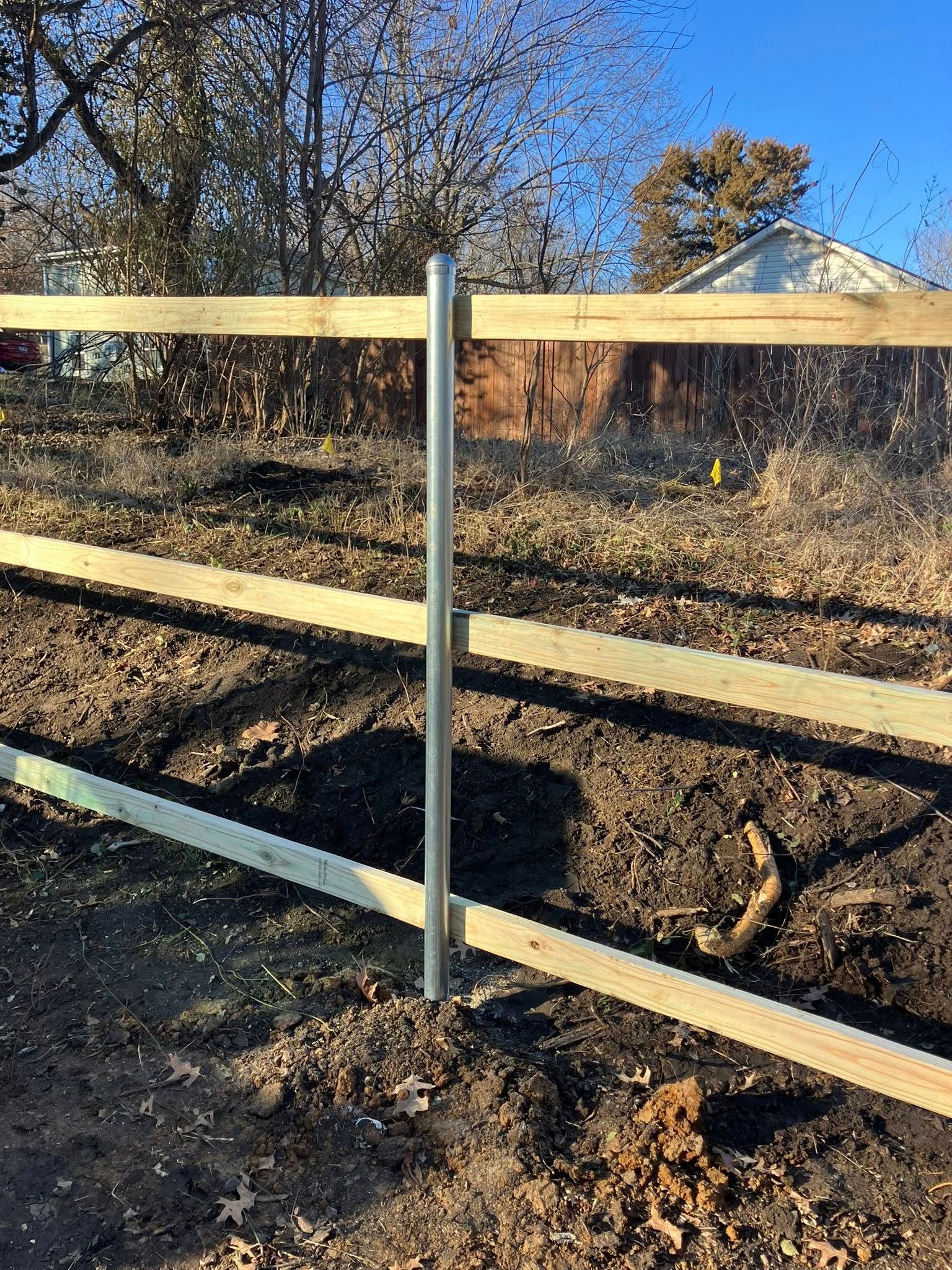 A metal fence post stands centered, supporting three horizontal wooden rails against a backdrop of bare trees and soil.