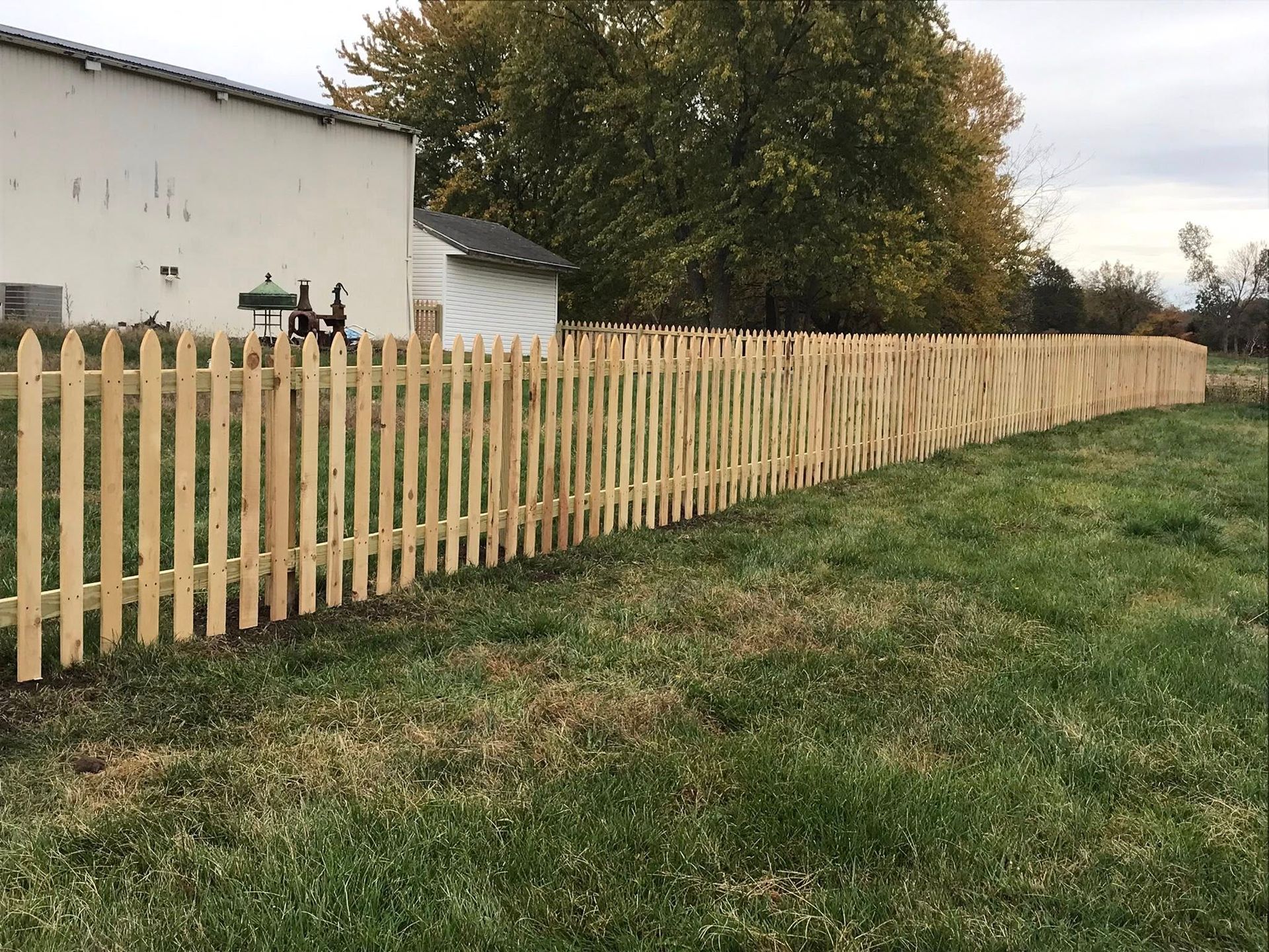 A wooden picket fence stretches across a green lawn in front of a white building and trees under a cloudy sky.