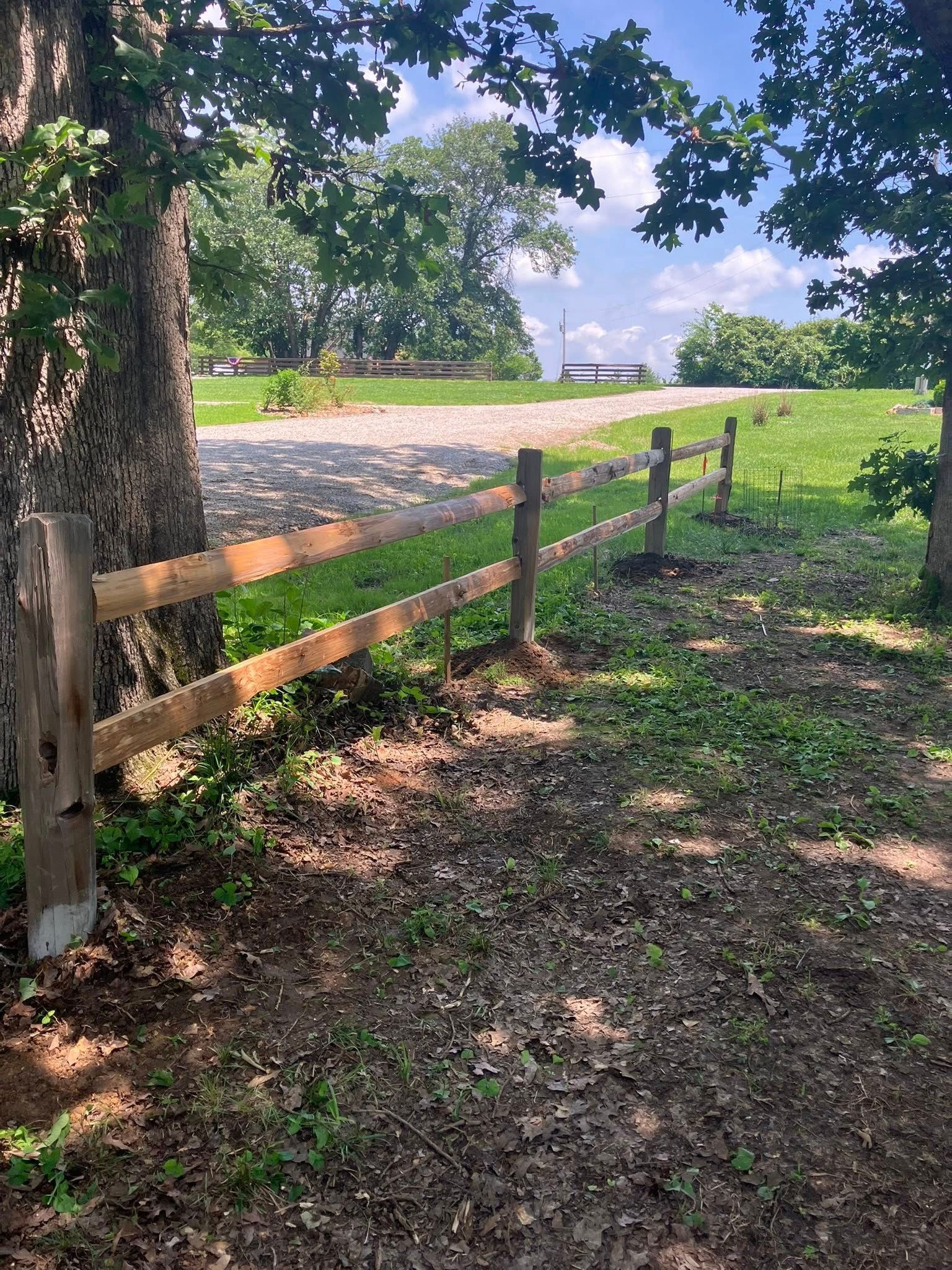 A wooden split-rail fence runs along a gravel path through a green, tree-lined meadow under a blue sky.