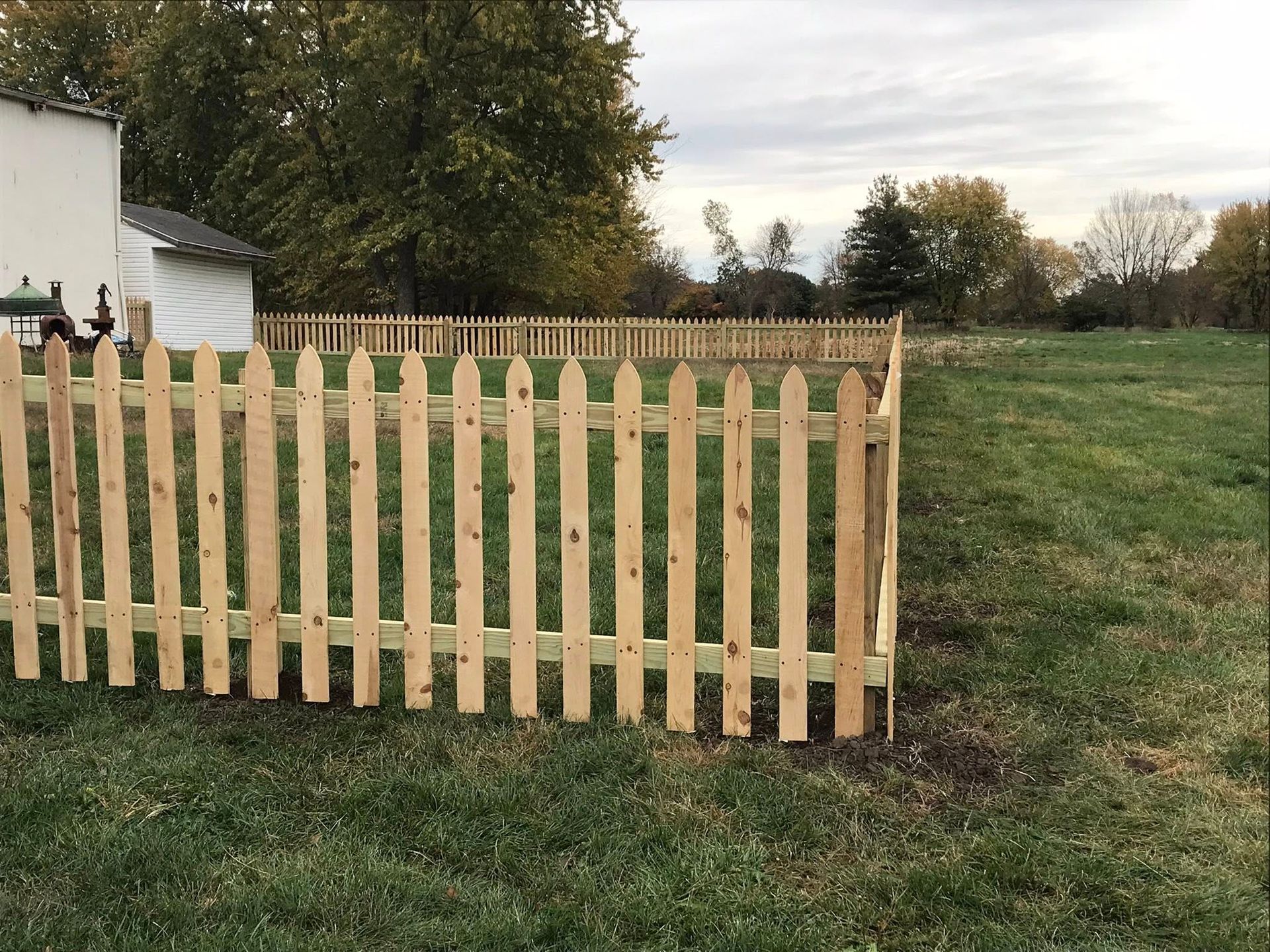 A newly constructed light-wood picket fence stands in a grassy field next to a house under a cloudy sky.