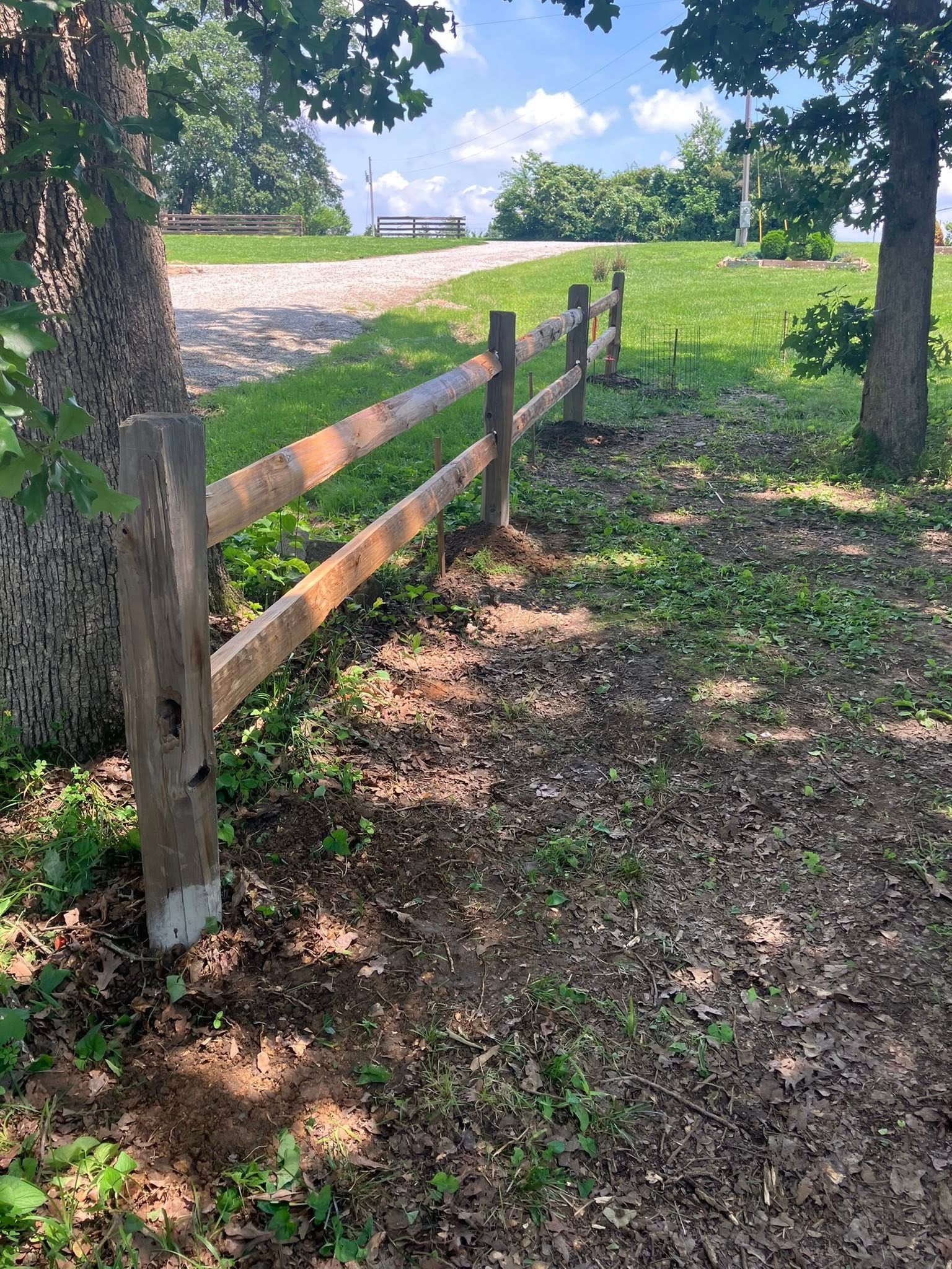 A weathered wooden post-and-rail fence runs along a grassy, tree-lined path under a bright blue sky.