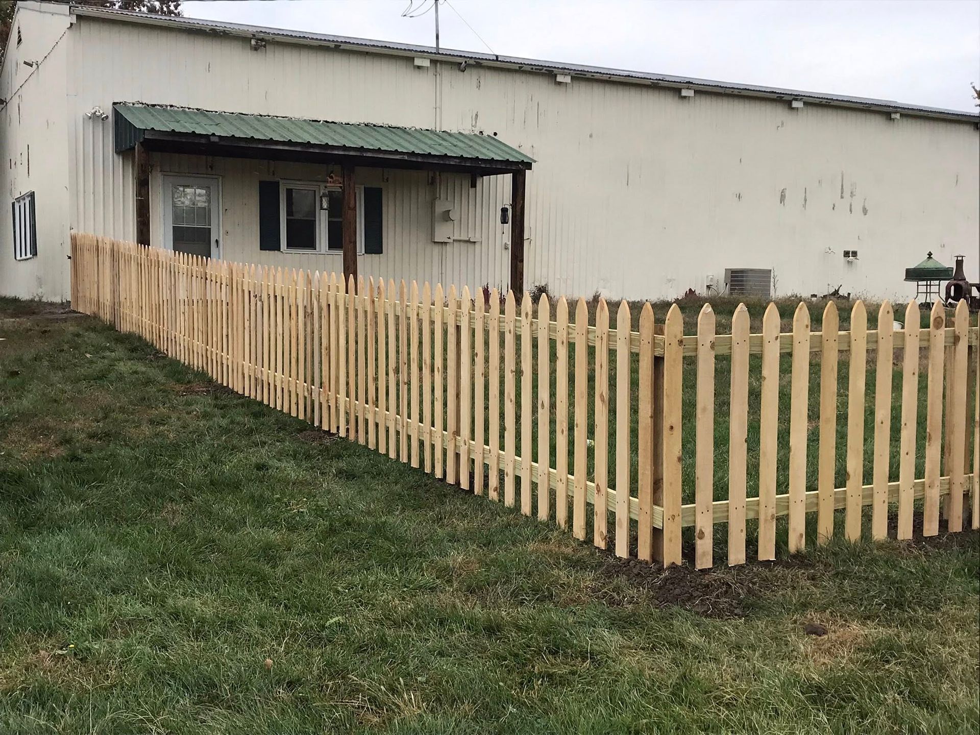 A new wooden picket fence encloses a grassy lawn in front of a white building with a small, green-roofed porch.