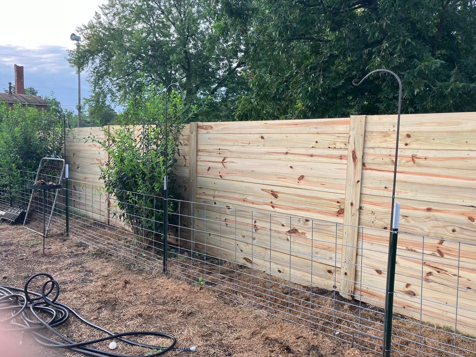A new horizontal wood fence panel with a wire mesh barrier along the bottom, set against trees and mulch.