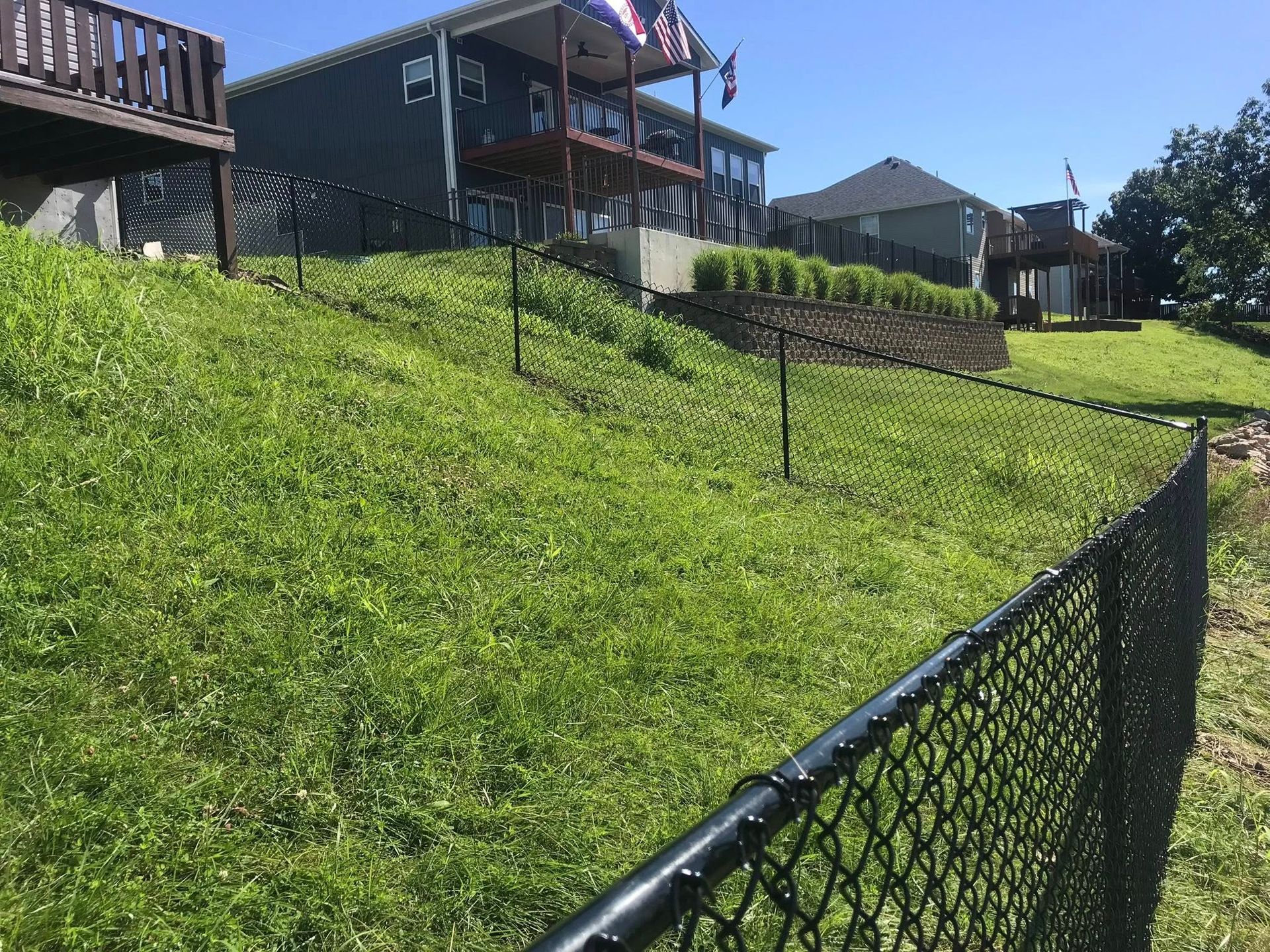 A grassy, sloped residential backyard with a black chain-link fence in the foreground and two houses in the background.