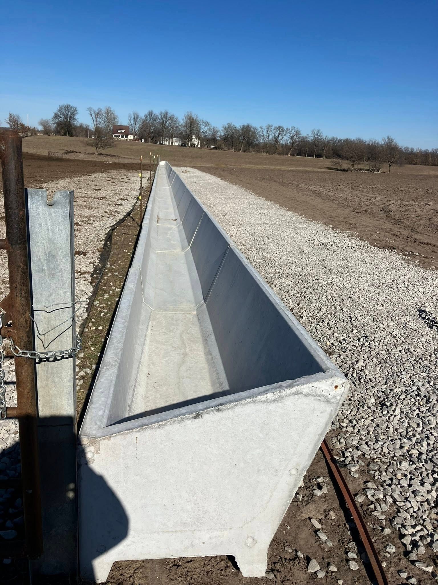 A long, narrow concrete livestock feed trough sitting on a bed of gravel in an open field.