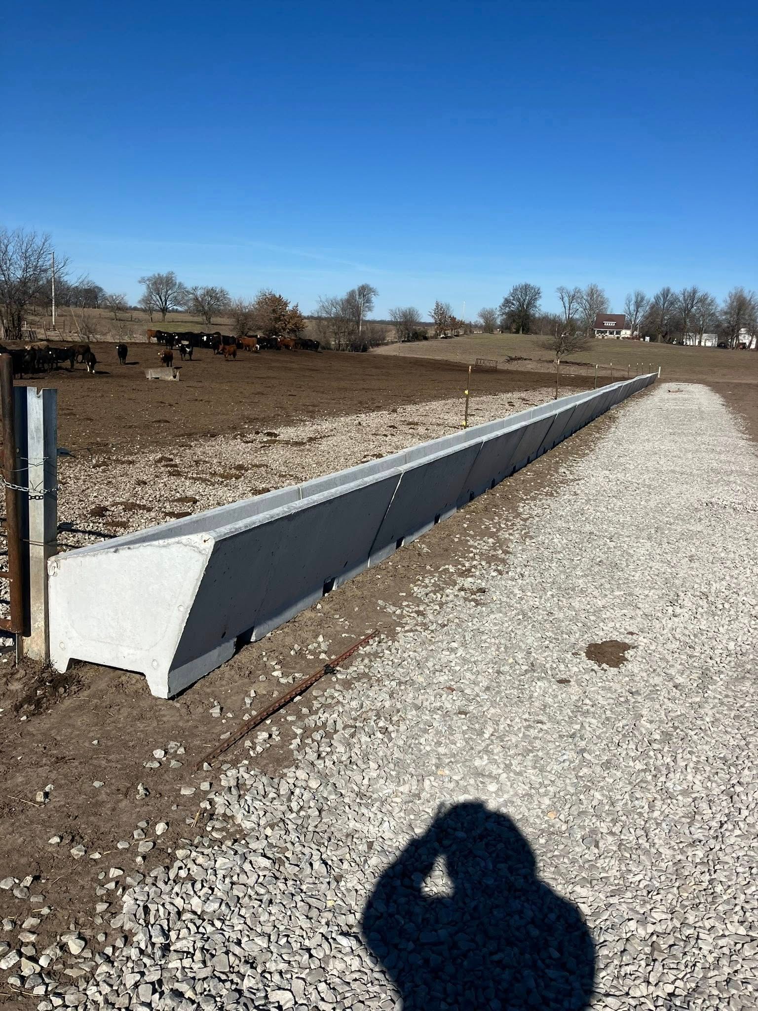 A long, concrete livestock feed bunk sitting between a gravel access path and a dirt pasture under a clear blue sky.