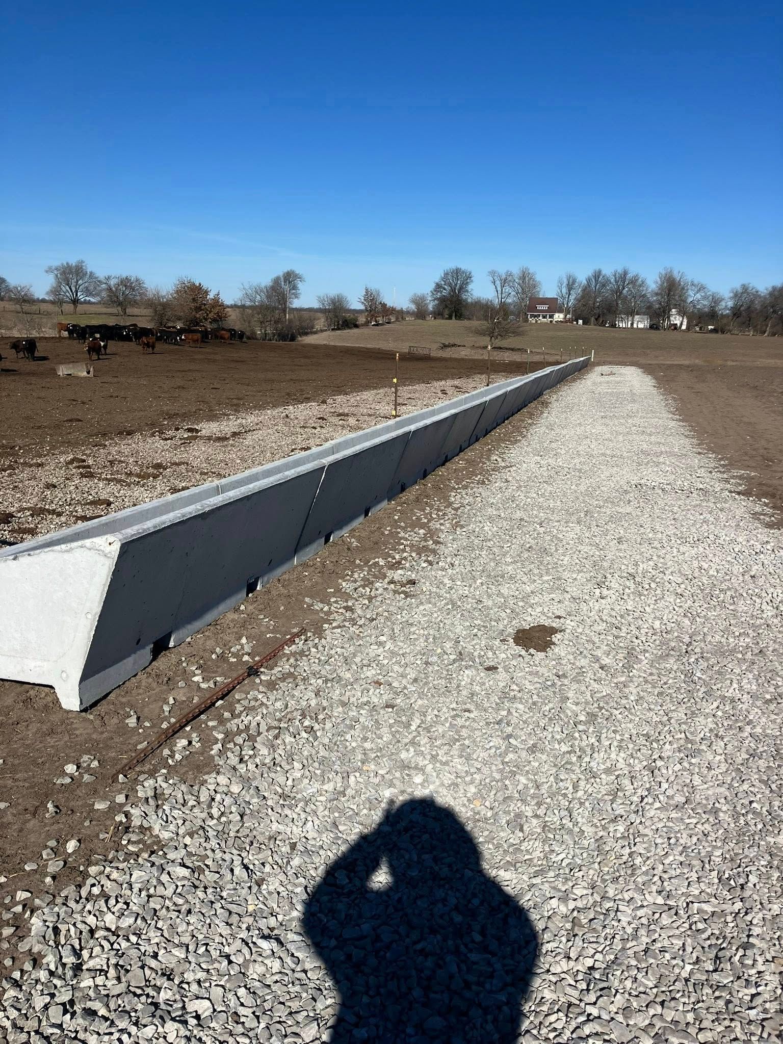 A long, concrete feed bunk sits beside a gravel path in a fenced pasture under a clear blue sky.