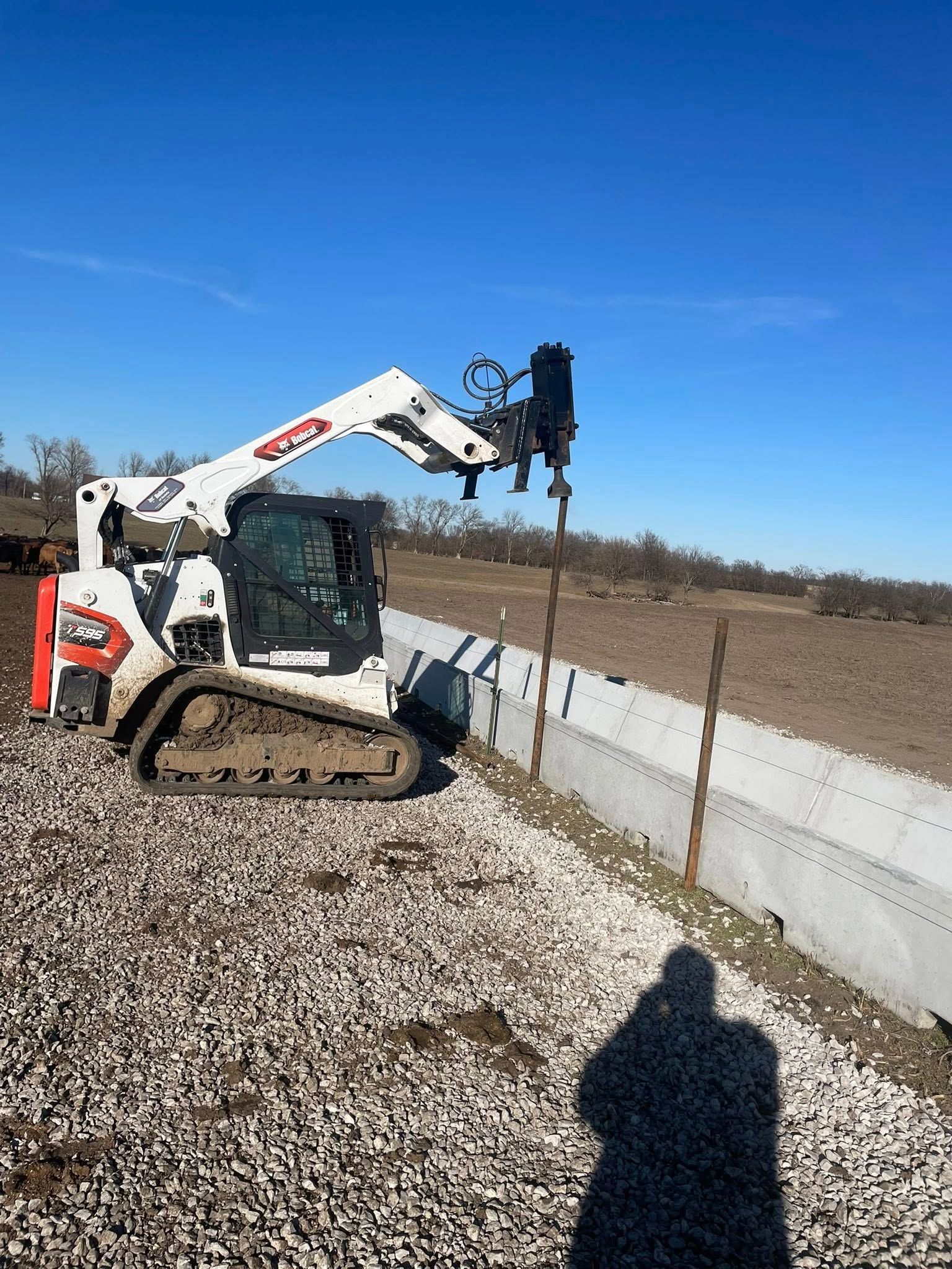 A white Bobcat compact track loader with a post driver attachment installing fence posts along a concrete barrier.