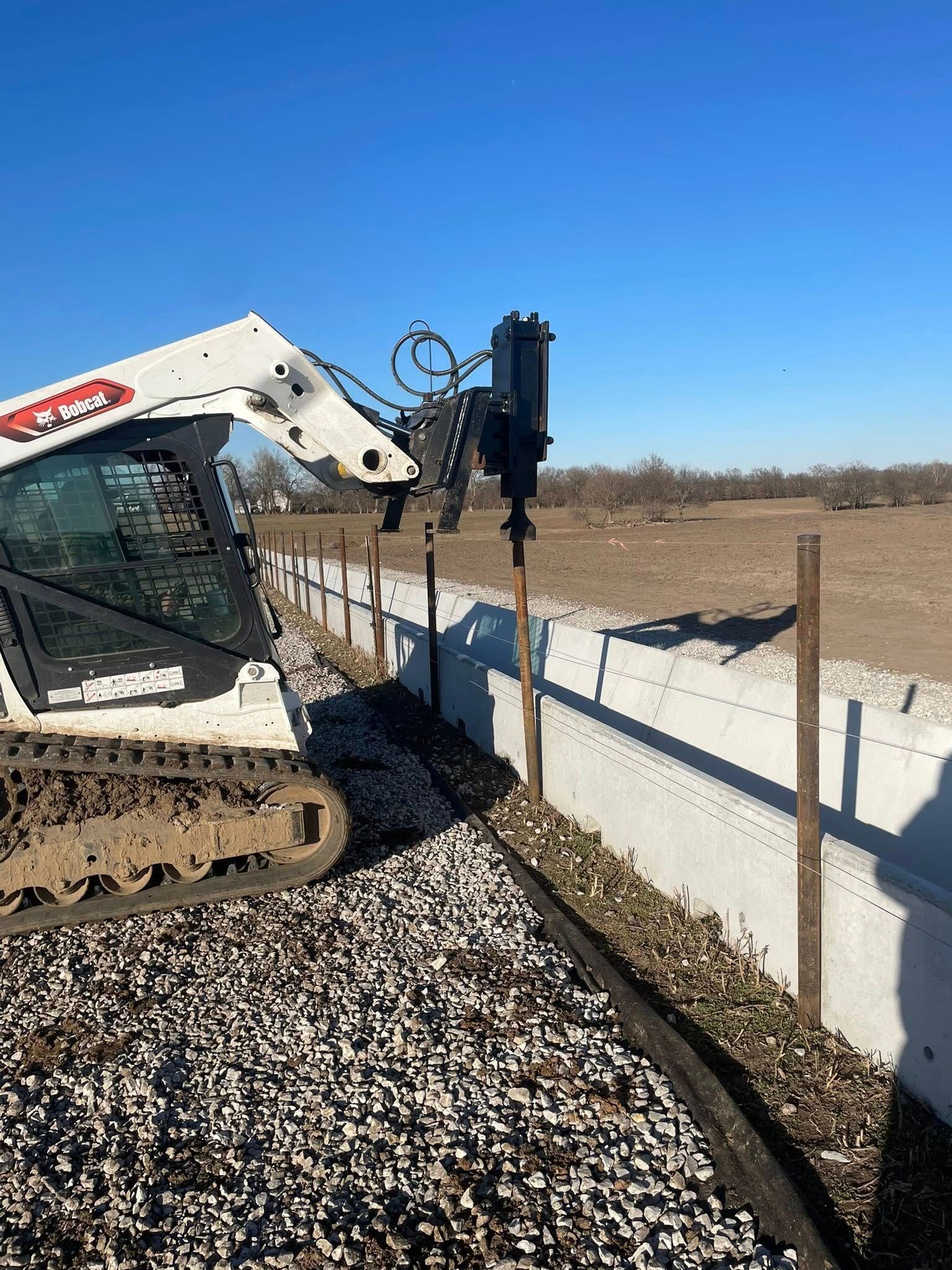 A white Bobcat skid-steer loader uses a hydraulic post driver to install metal fence posts along a concrete barrier.