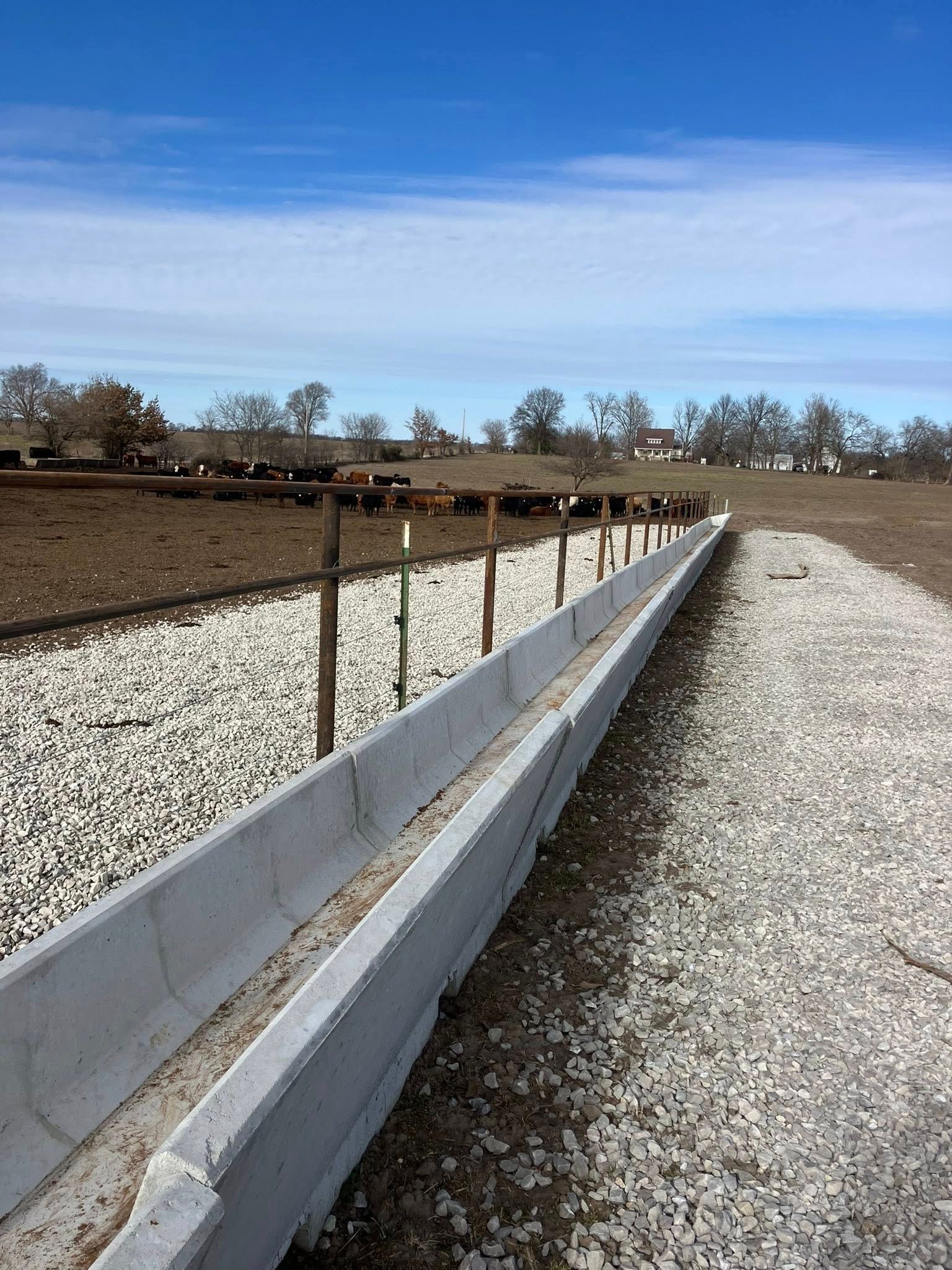 A long concrete livestock feeding trough runs beside a gravel path and wooden fence in a pasture with cattle in the distance.