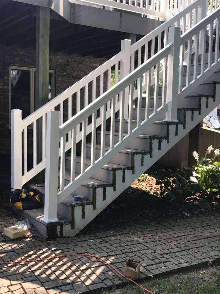 A set of outdoor stairs with white railings and vertical balusters leading up to a deck over a paved patio area.