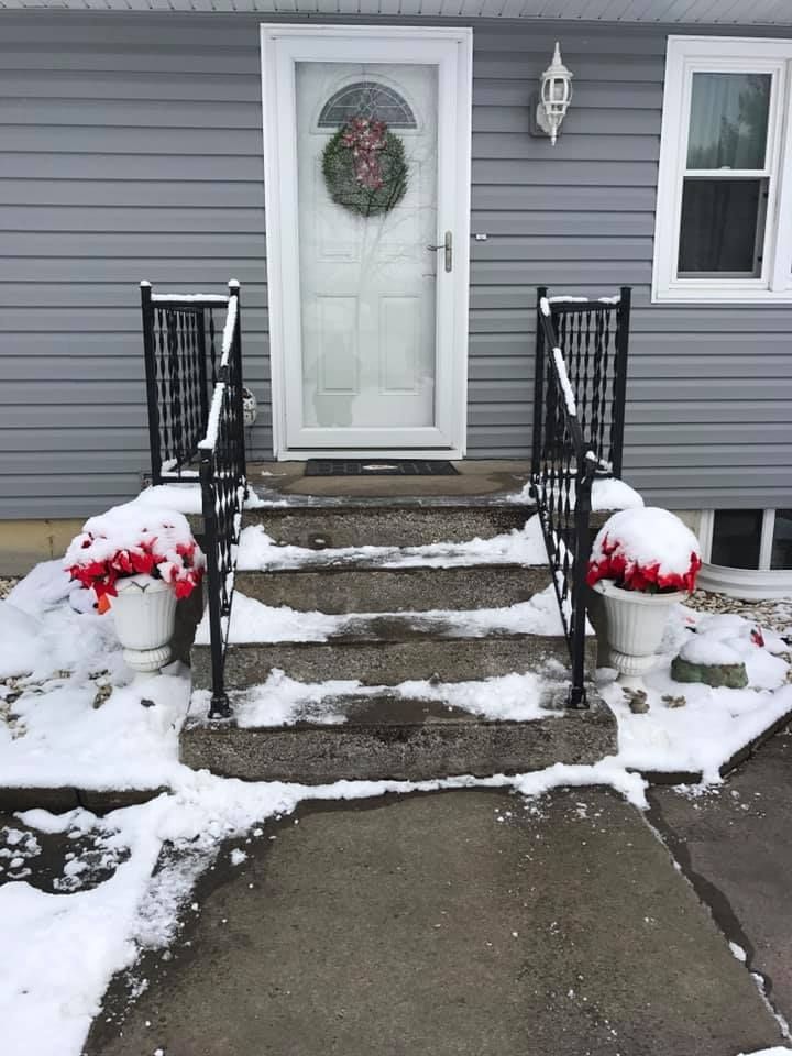 Concrete front steps with black railings, decorated with two pots of red flowers, leading to a white door with a wreath.
