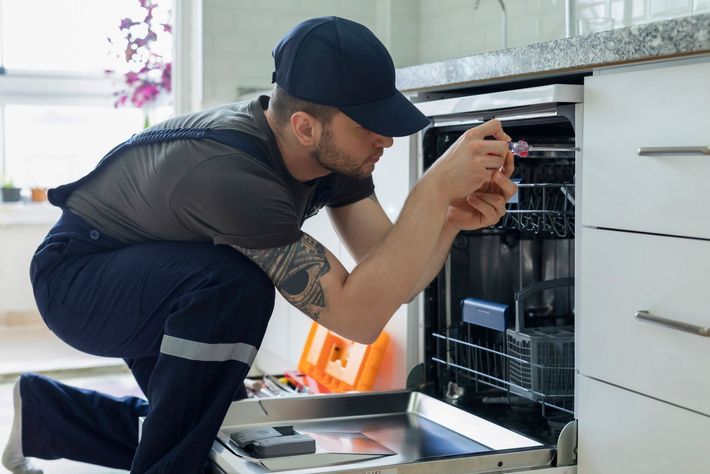 A repairman in a cap and overalls fixes a dishwasher in a kitchen, using a screwdriver.