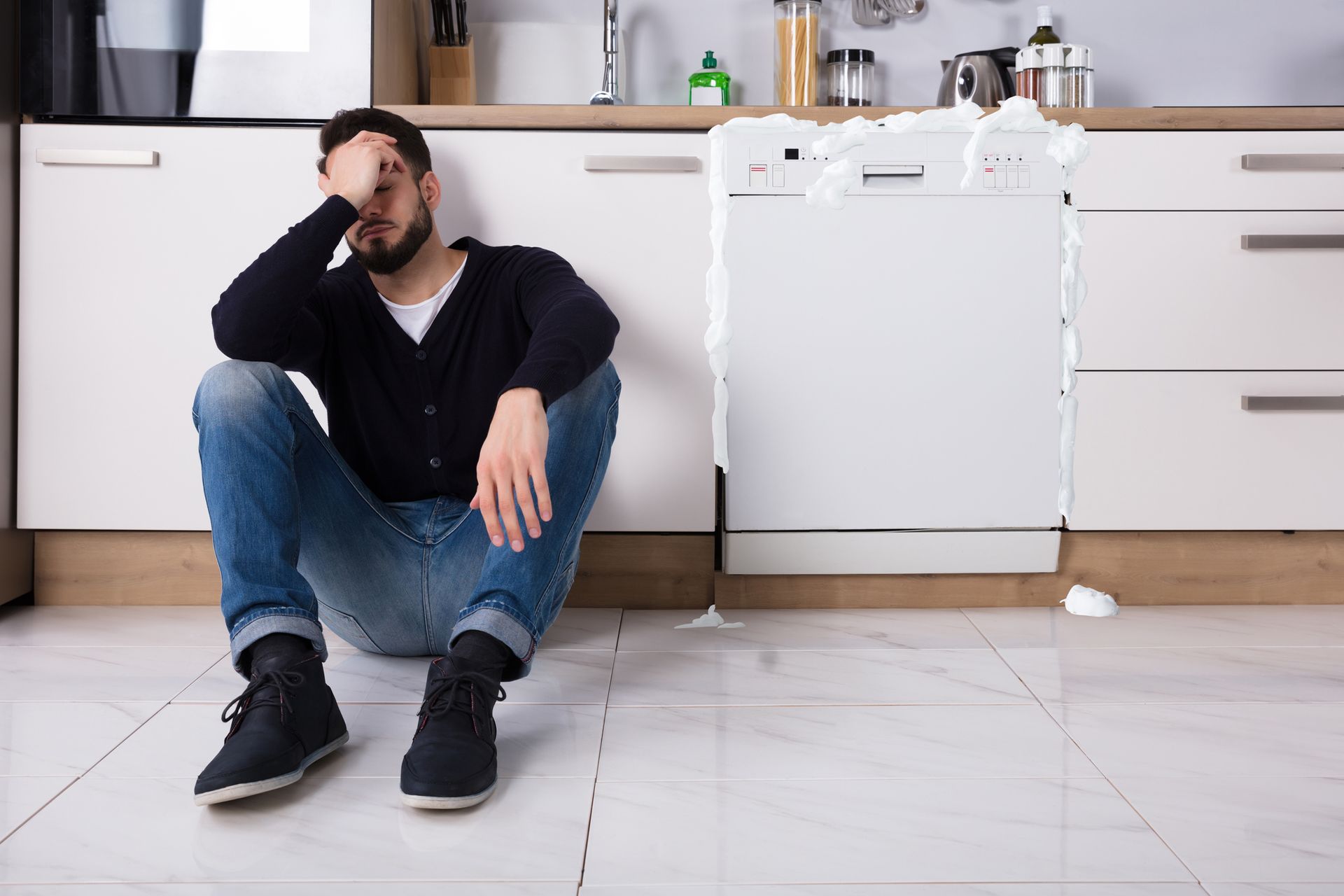 Man sits on the kitchen floor, looking stressed, with foam overflowing from an open dishwasher.