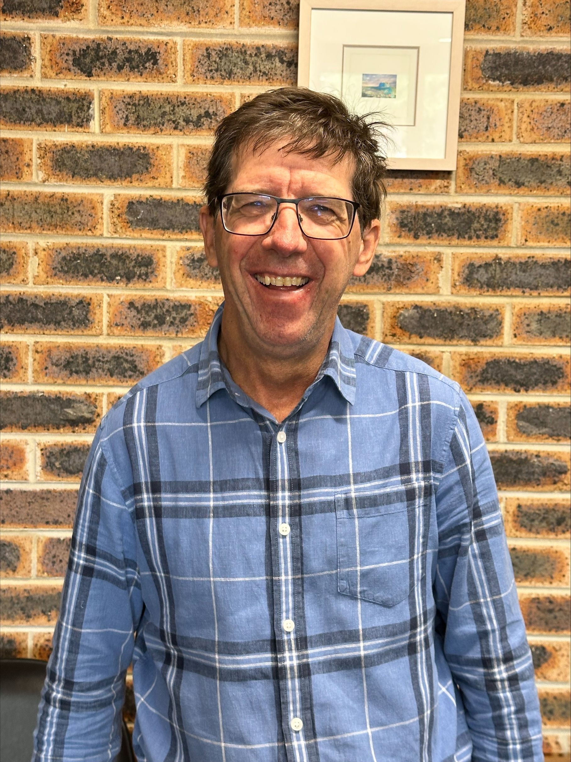 A man wearing glasses and a plaid shirt is smiling in front of a brick wall.