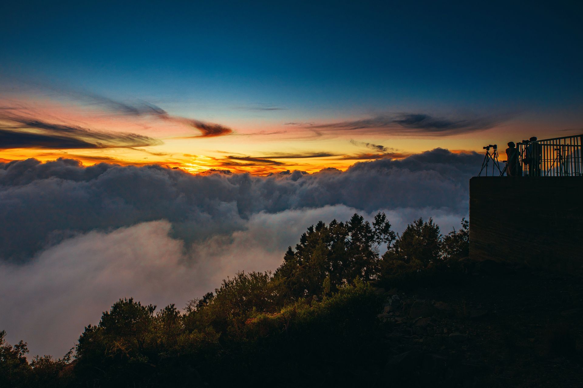 Sunset over a sea of clouds from a hilltop viewing platform with silhouetted figures.