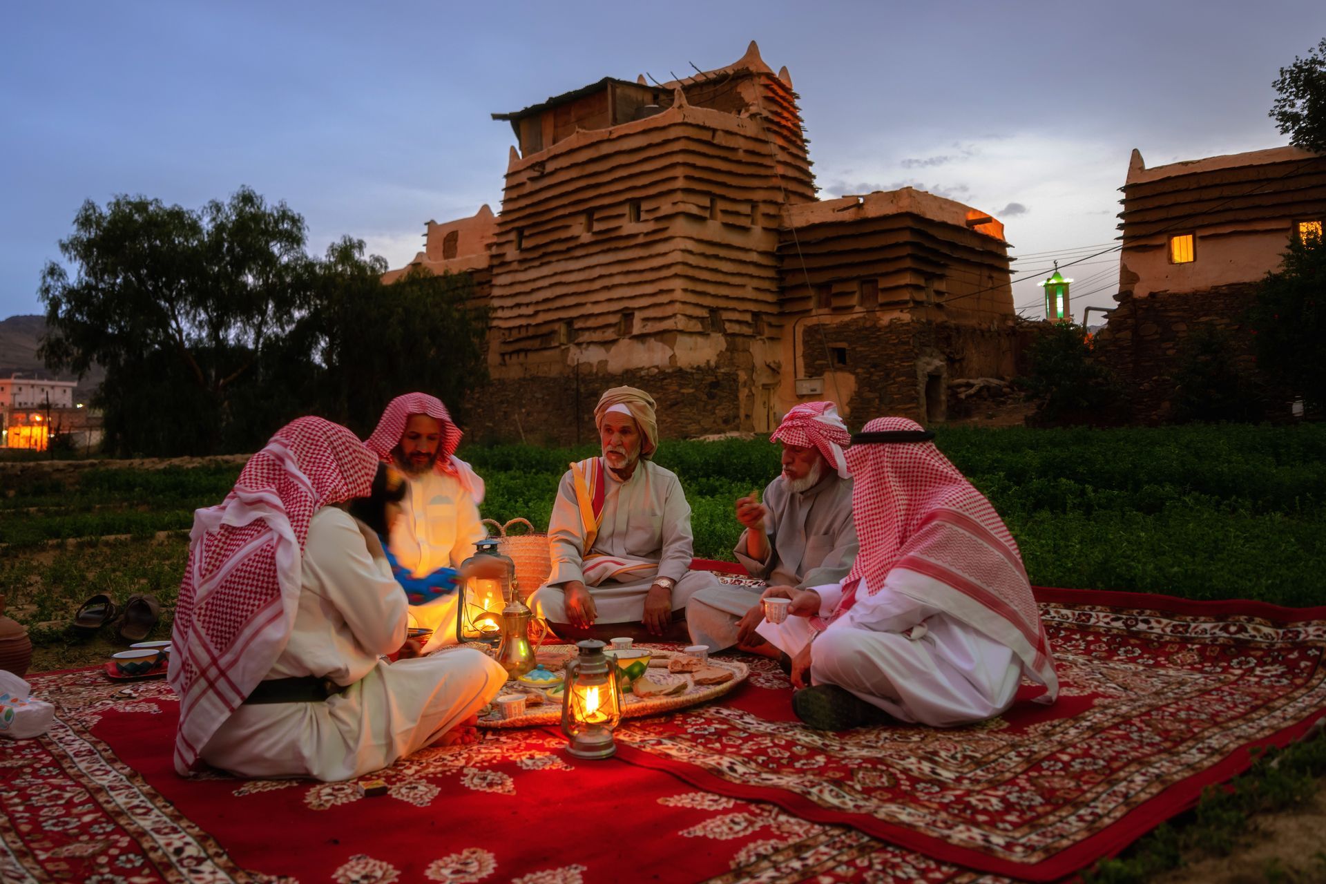 Men in traditional attire sit on a red rug, eating near a mud-brick building at dusk. Lanterns provide light.