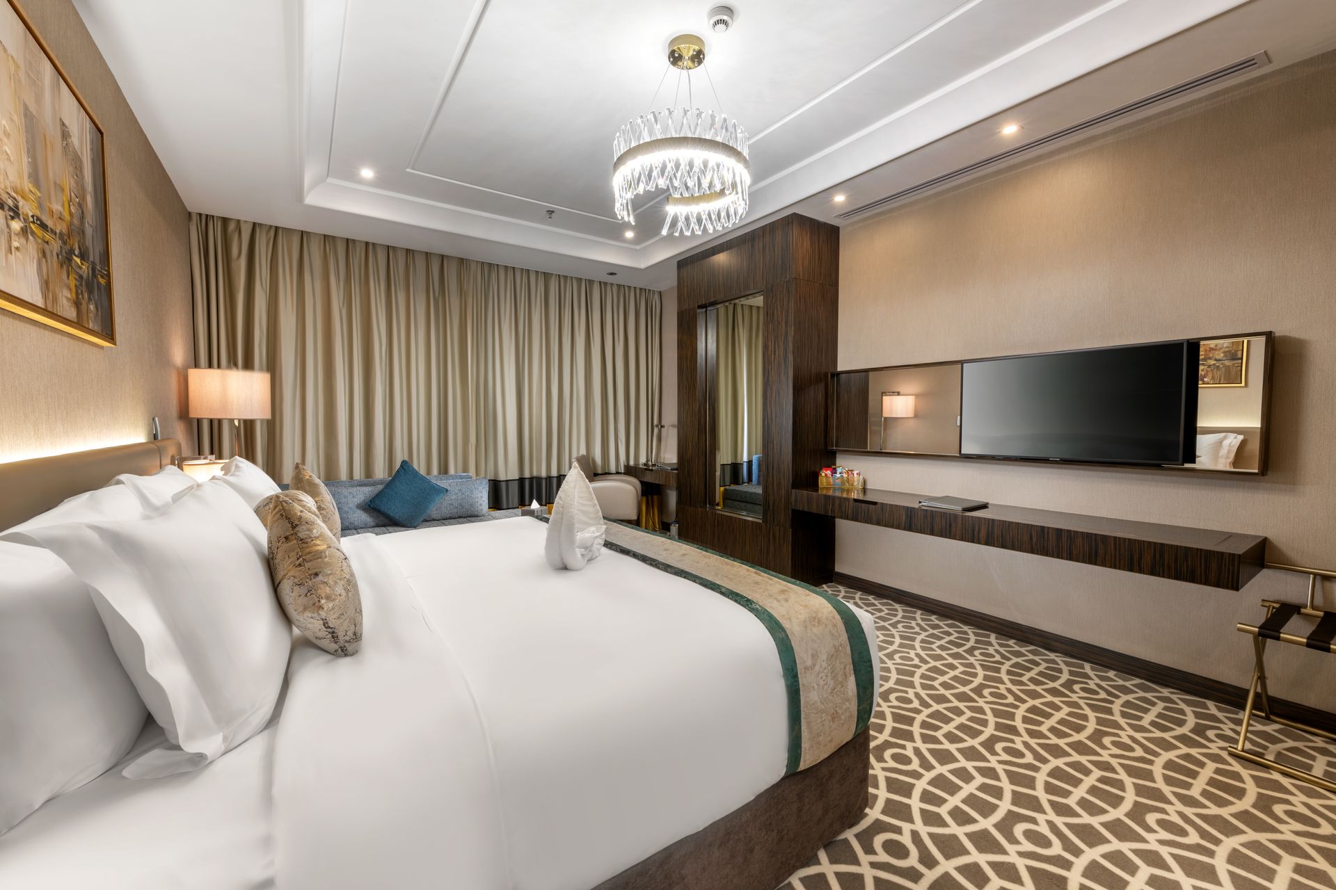 Hotel room with king bed, TV, desk, and chandelier. Beige and brown tones.