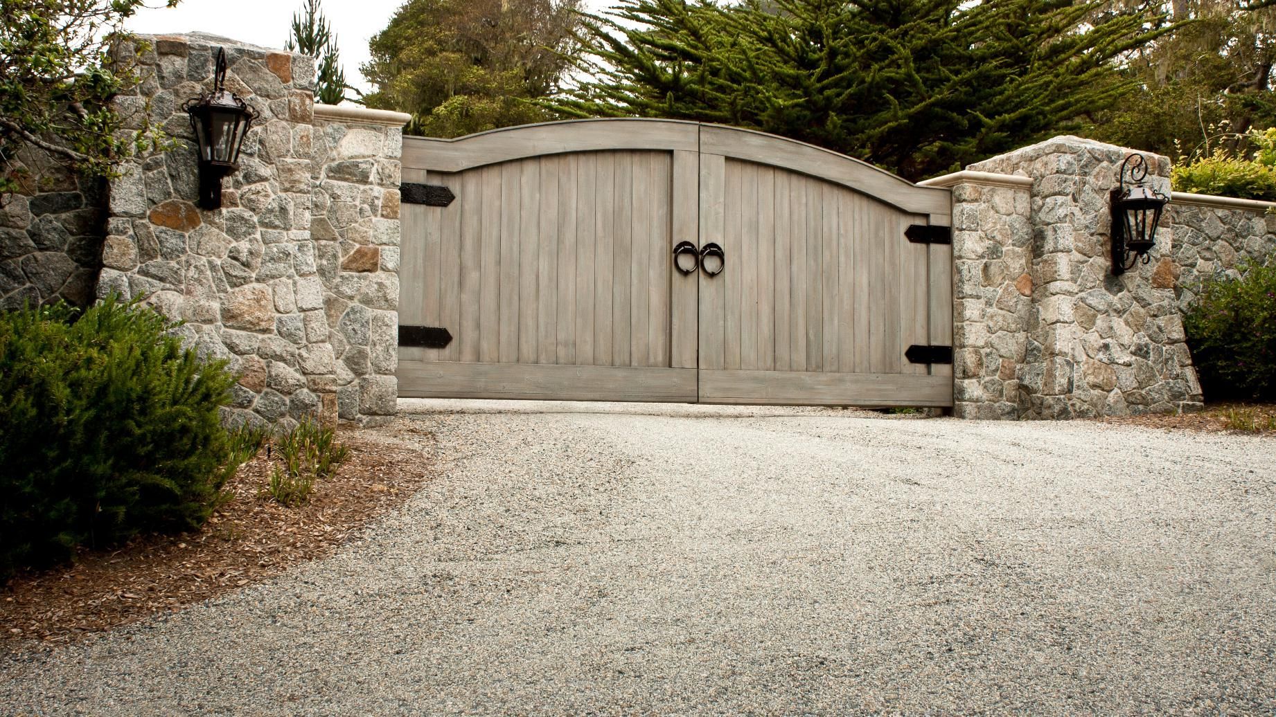 A wooden gate is surrounded by a stone wall and gravel driveway.