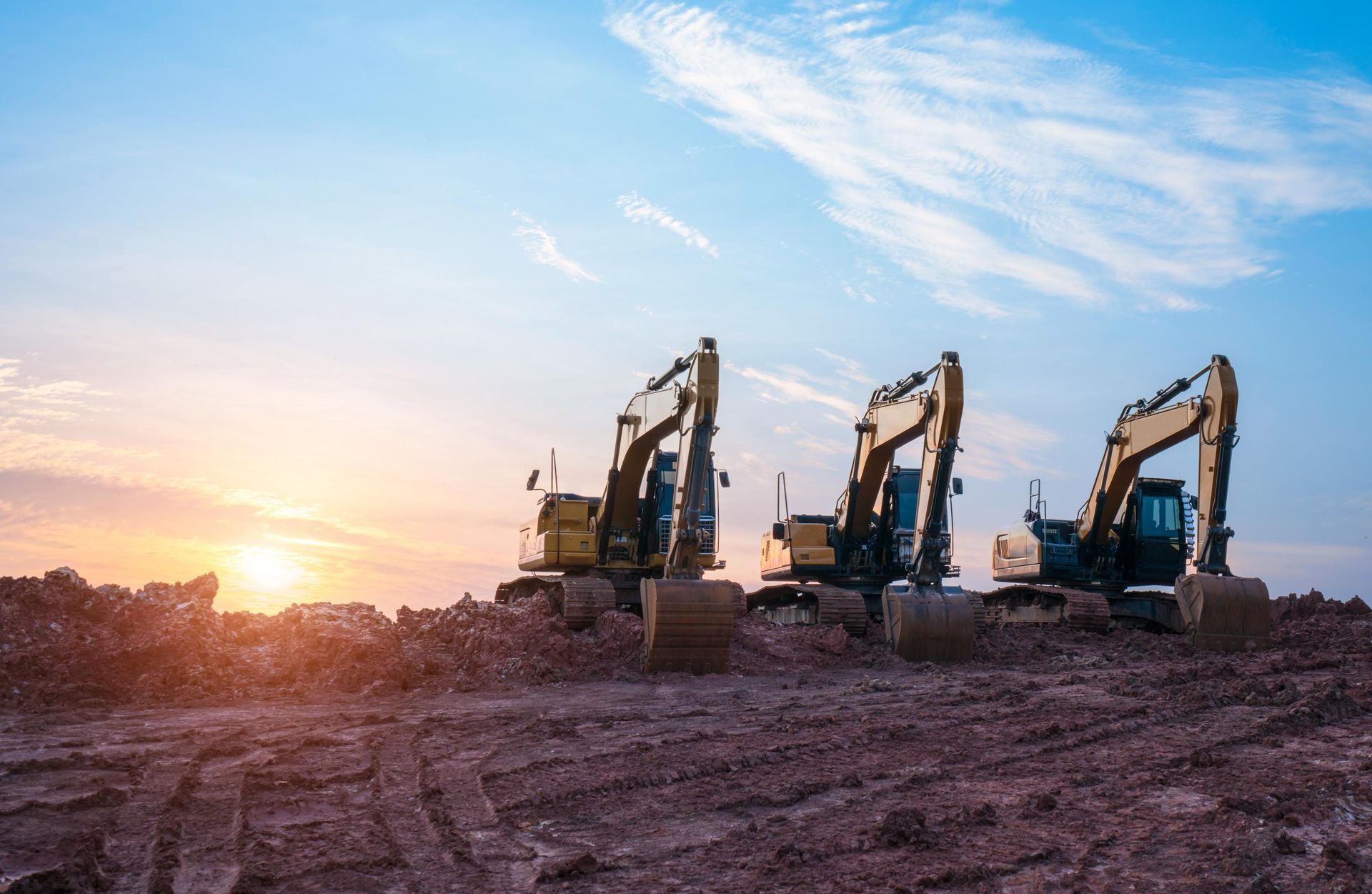 A row of construction vehicles are parked in a dirt field at sunset.