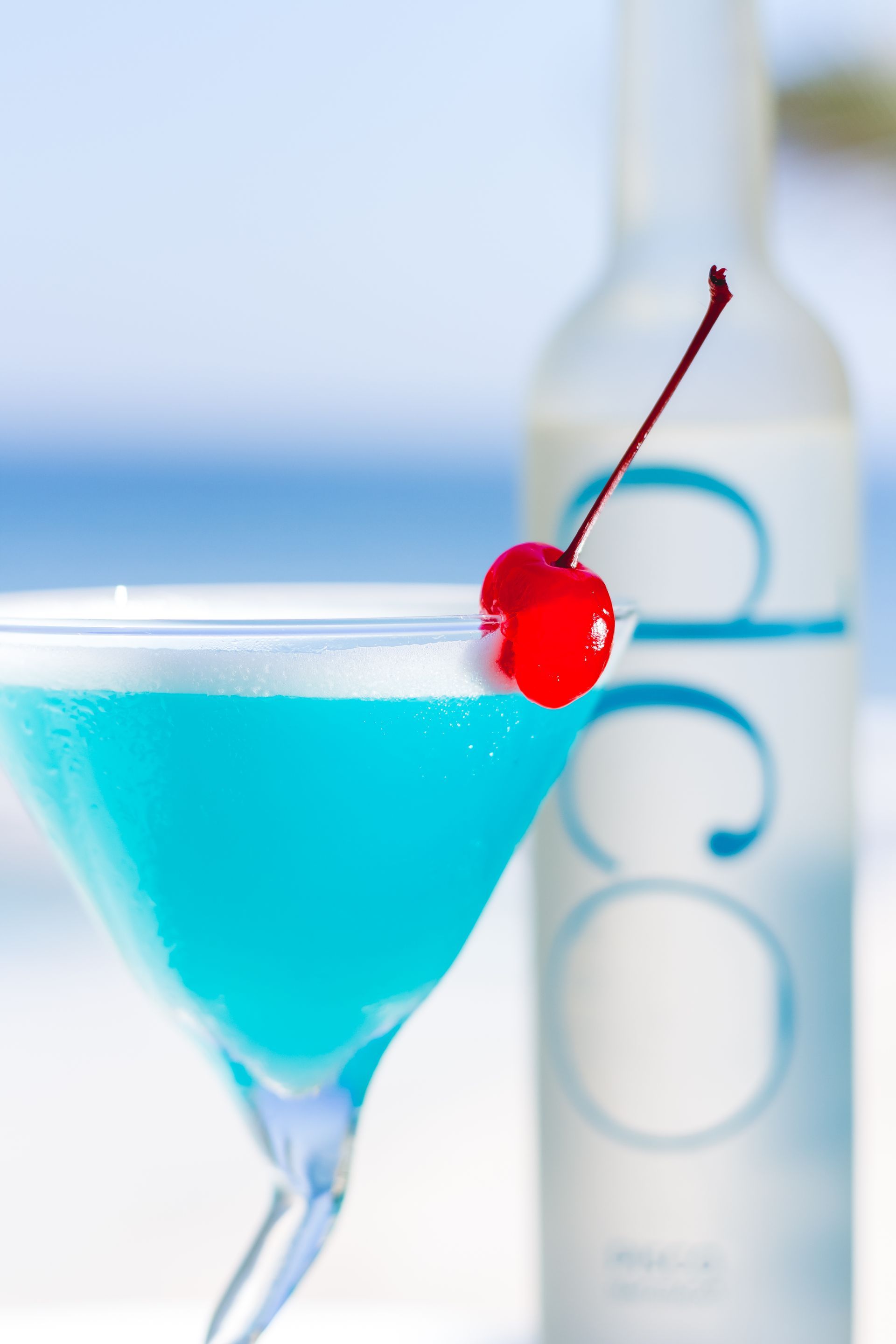A blue martini with a cherry on top next to a bottle of coco