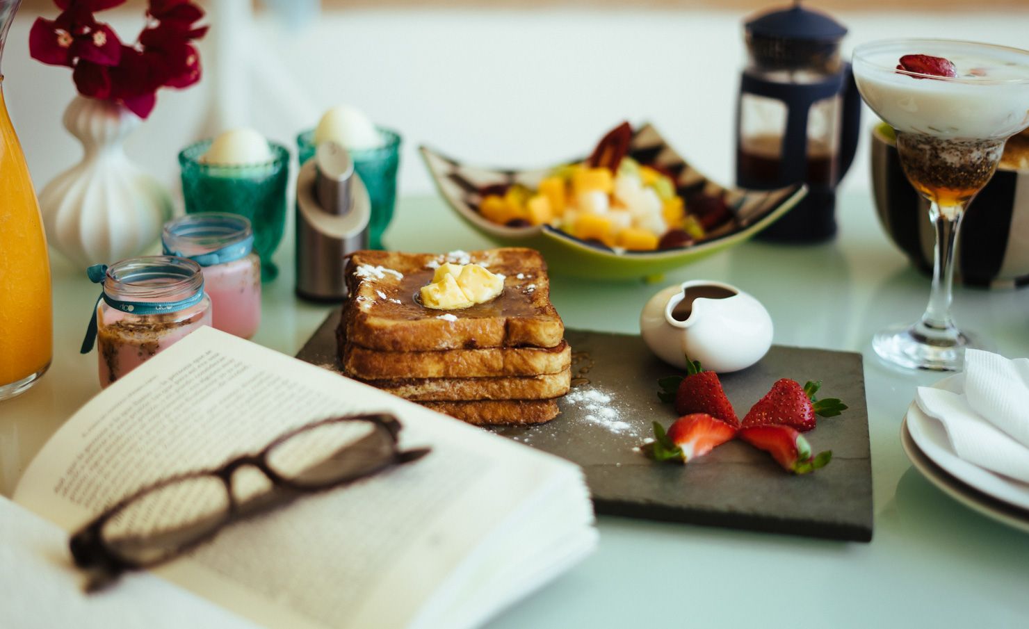 A table topped with a book , glasses , toast , fruit , and drinks.