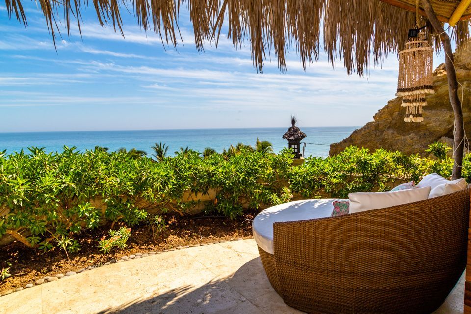 A wicker bed is sitting under a thatched roof overlooking the ocean.