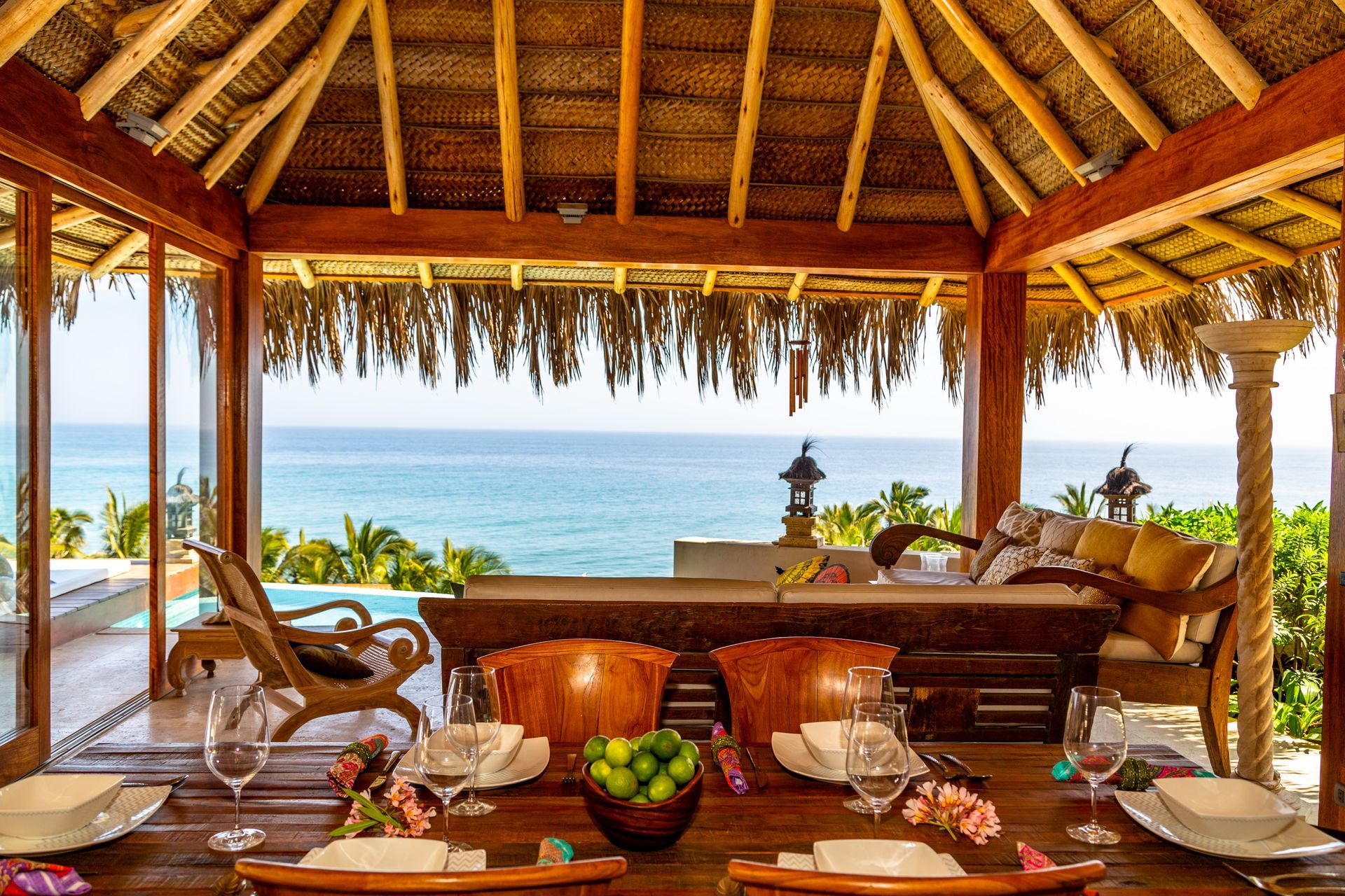 A table with plates and glasses on it in front of a thatched roof overlooking the ocean.