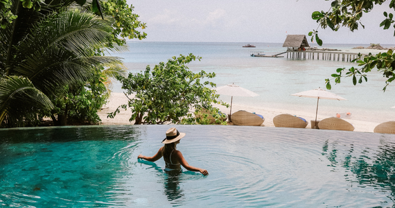 Woman in a Turquoise Infinity Pool, Ocean View With Beach Umbrellas, Trees, and Small Hut — Midcoast Financial Planning Group in Tuncurry, NSW