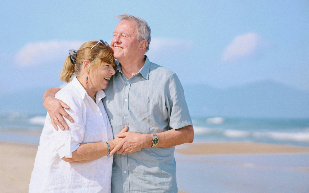 Couple Embracing on Beach, Smiling Man Has Arm Around Woman — Midcoast Financial Planning Group in Tuncurry, NSW