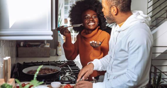 Couple Cooking Together in A Kitchen. Woman Tastes Food — Midcoast Financial Planning Group in Tuncurry, NSW