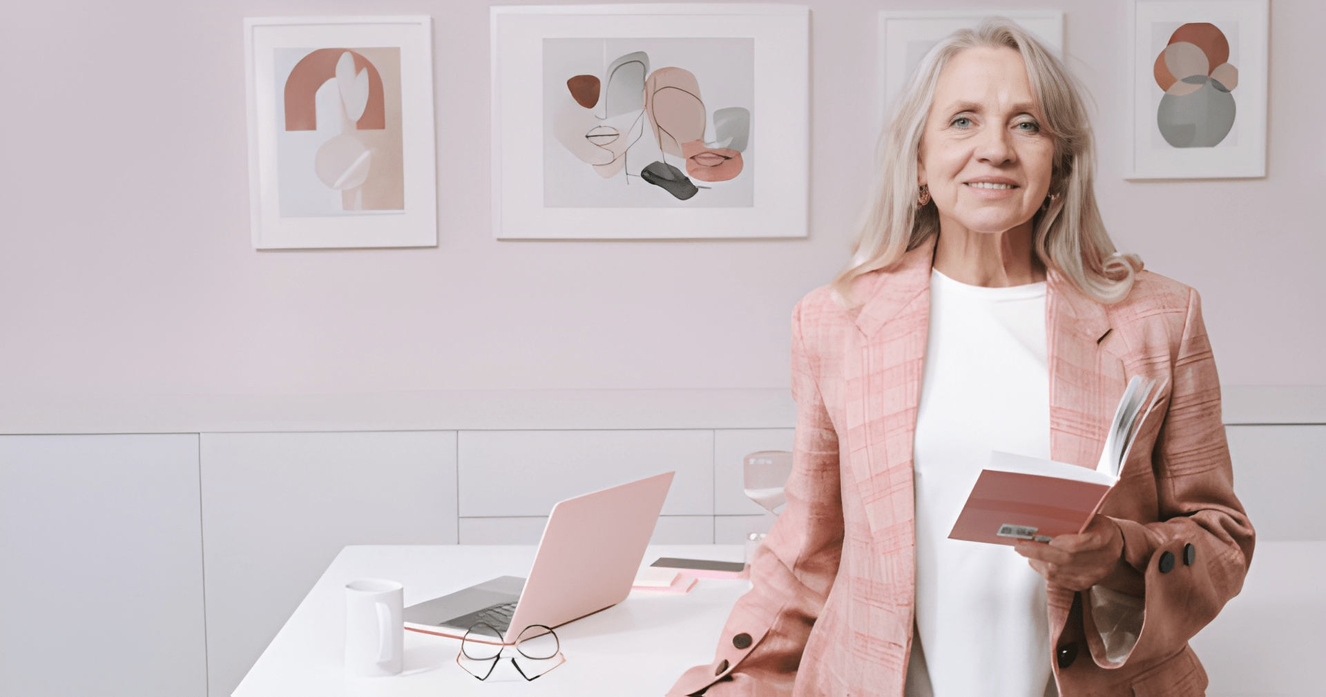 Woman in Pink Blazer Smiles, Holding Notebook Near a Desk With a Laptop and Artwork-filled Wall — Midcoast Financial Planning Group in Tuncurry, NSW