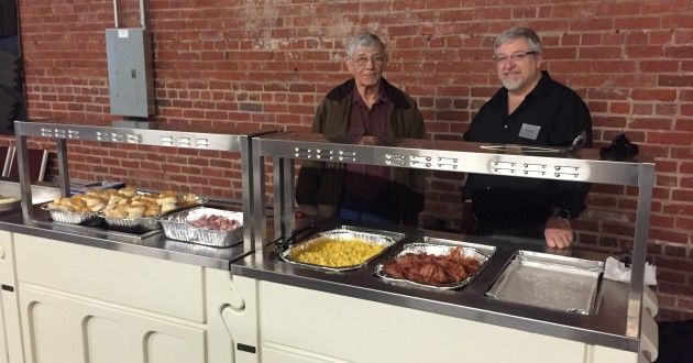 Two men are standing in front of a buffet line filled with food.