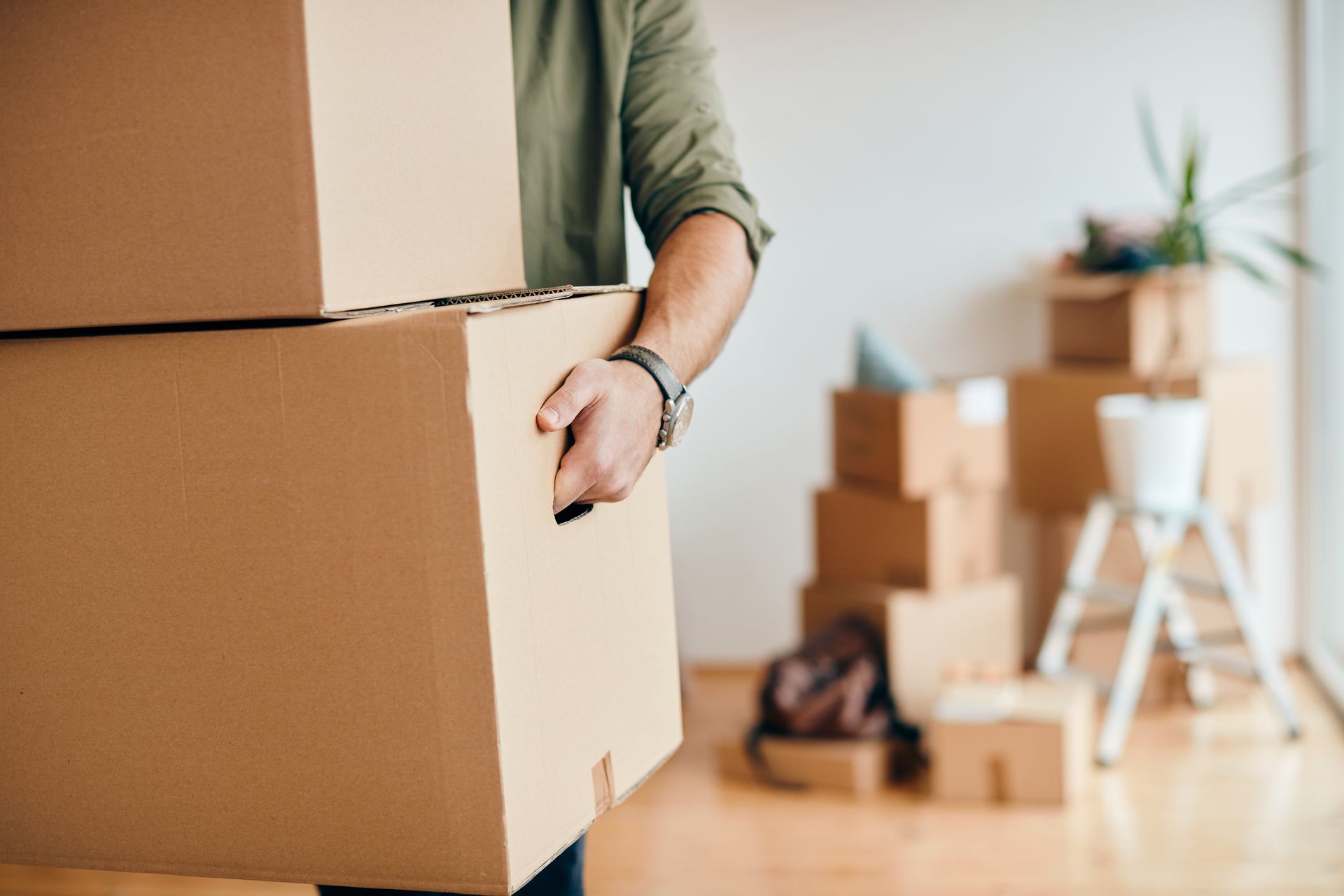 Person carrying stacked cardboard moving boxes in a bright room with more boxes in the background