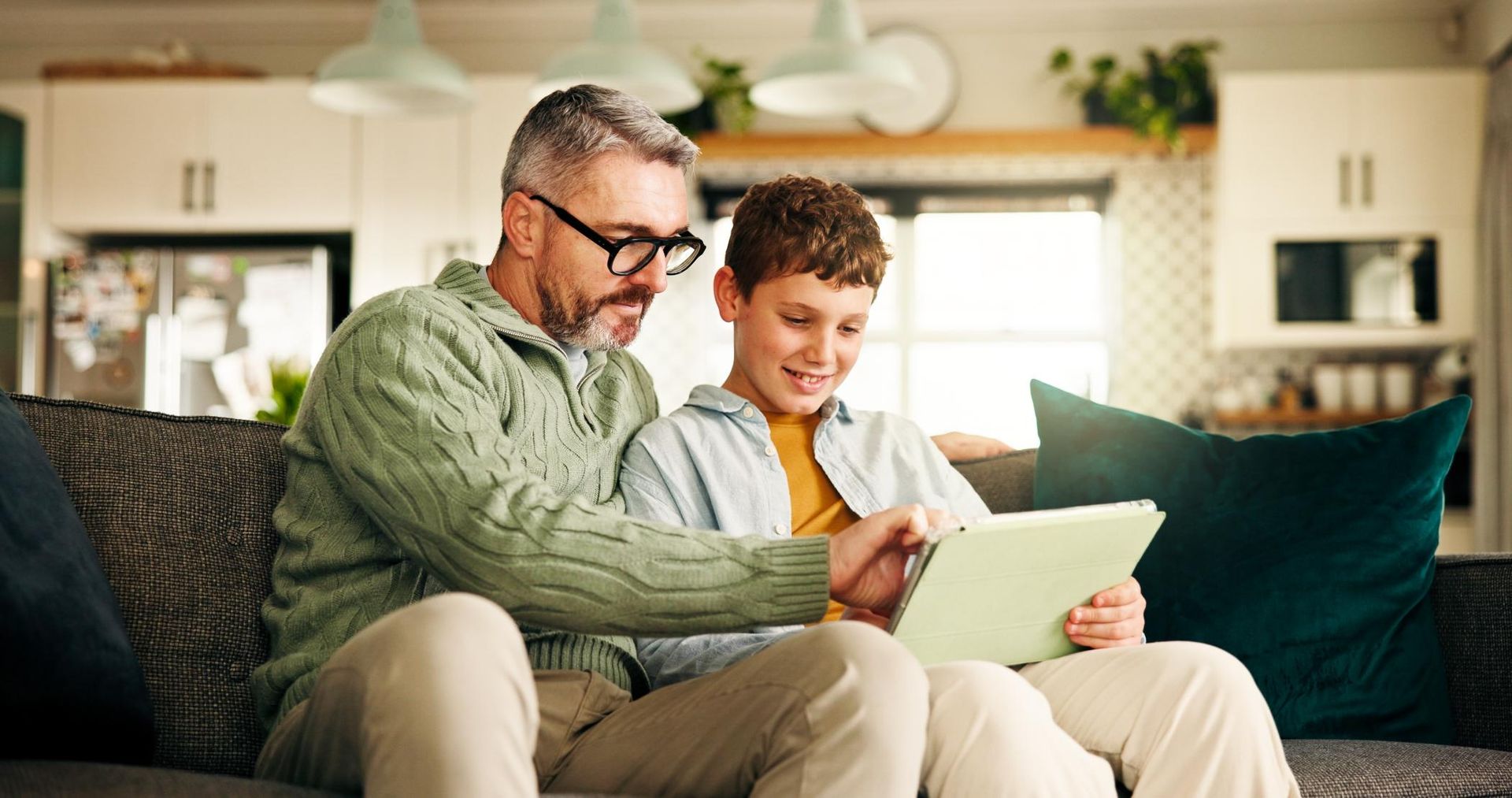 Man and child sit on a couch, reading together from a tablet in a bright living room.