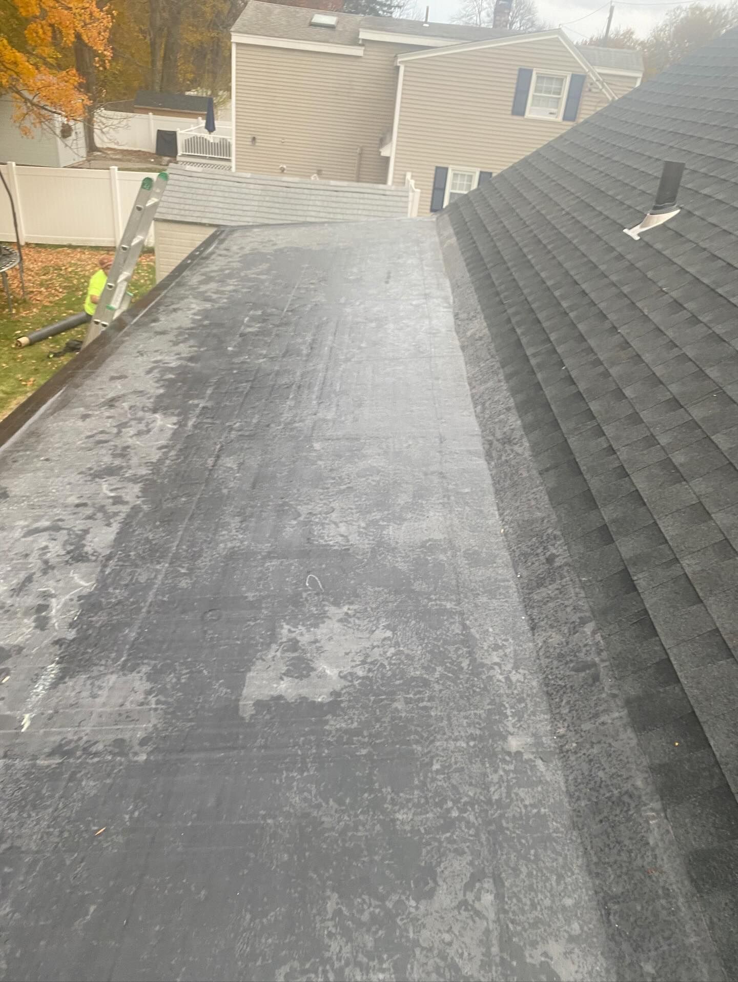 A view from a roof showing a flat, weathered black membrane section adjacent to a sloped asphalt shingle roof.
