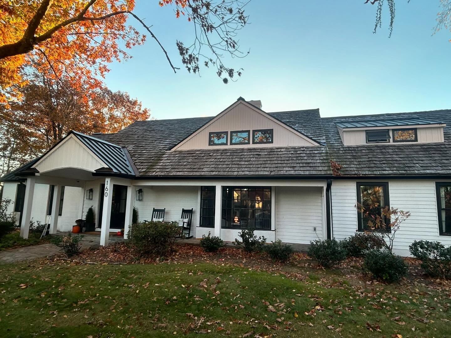 A white, single-story house with a wooden shingle roof and a front porch, surrounded by autumn trees on a sunny day.
