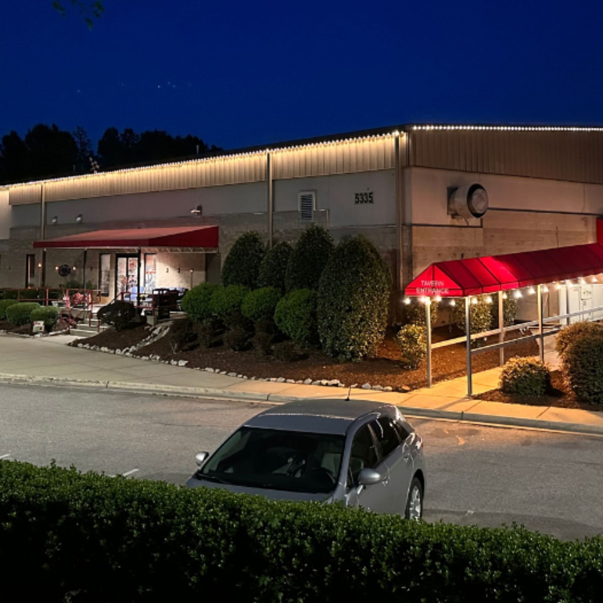 Silvers, Inc. - Nighttime exterior of a building with red awnings and string lights, a parked car, and bushes.