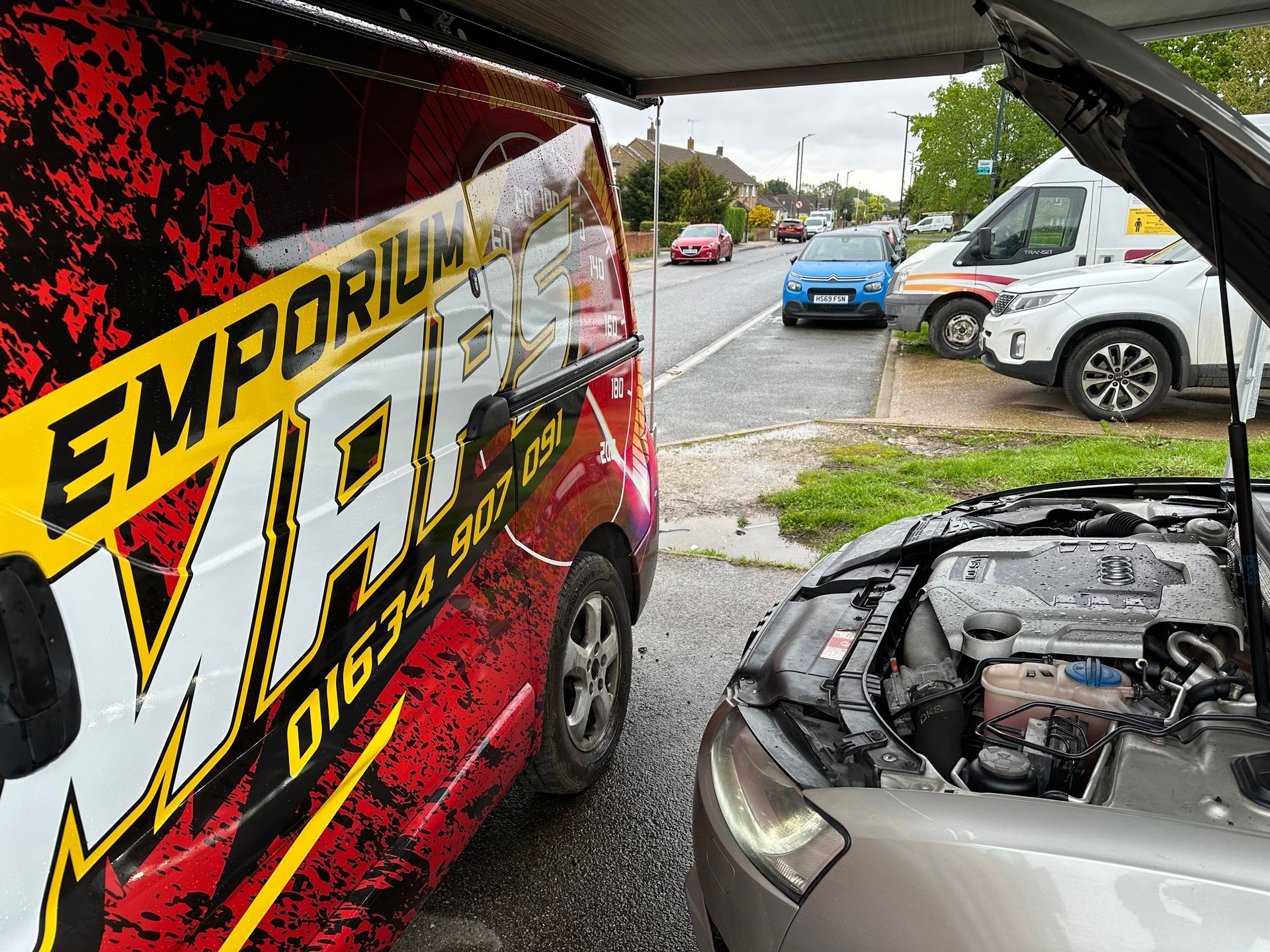 A red and white van with the word emporium on it is parked next to a car with its hood open.